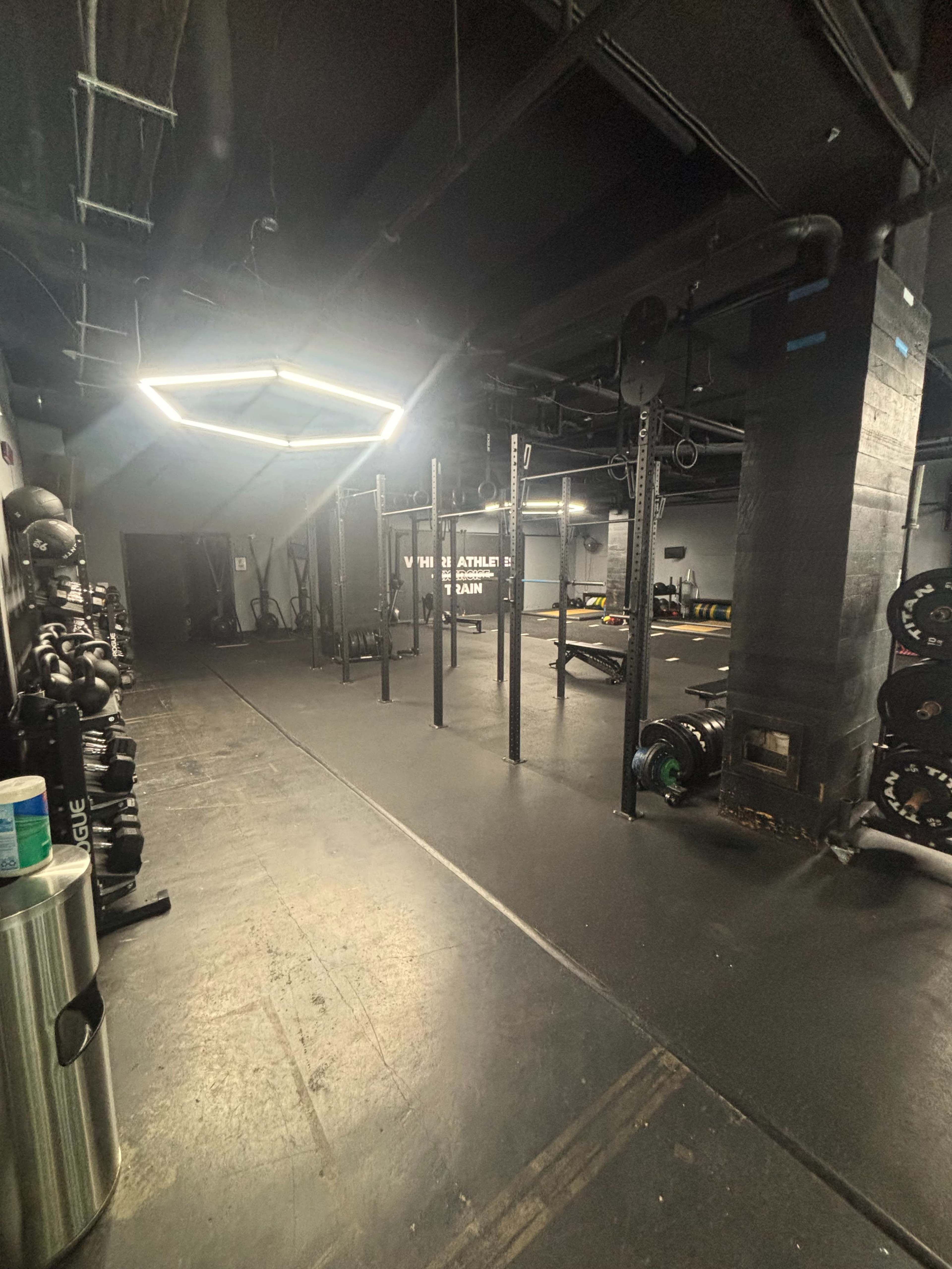 The image shows a dimly lit gym with weightlifting equipment, including benches, barbells, and kettlebells, arranged in an open space.