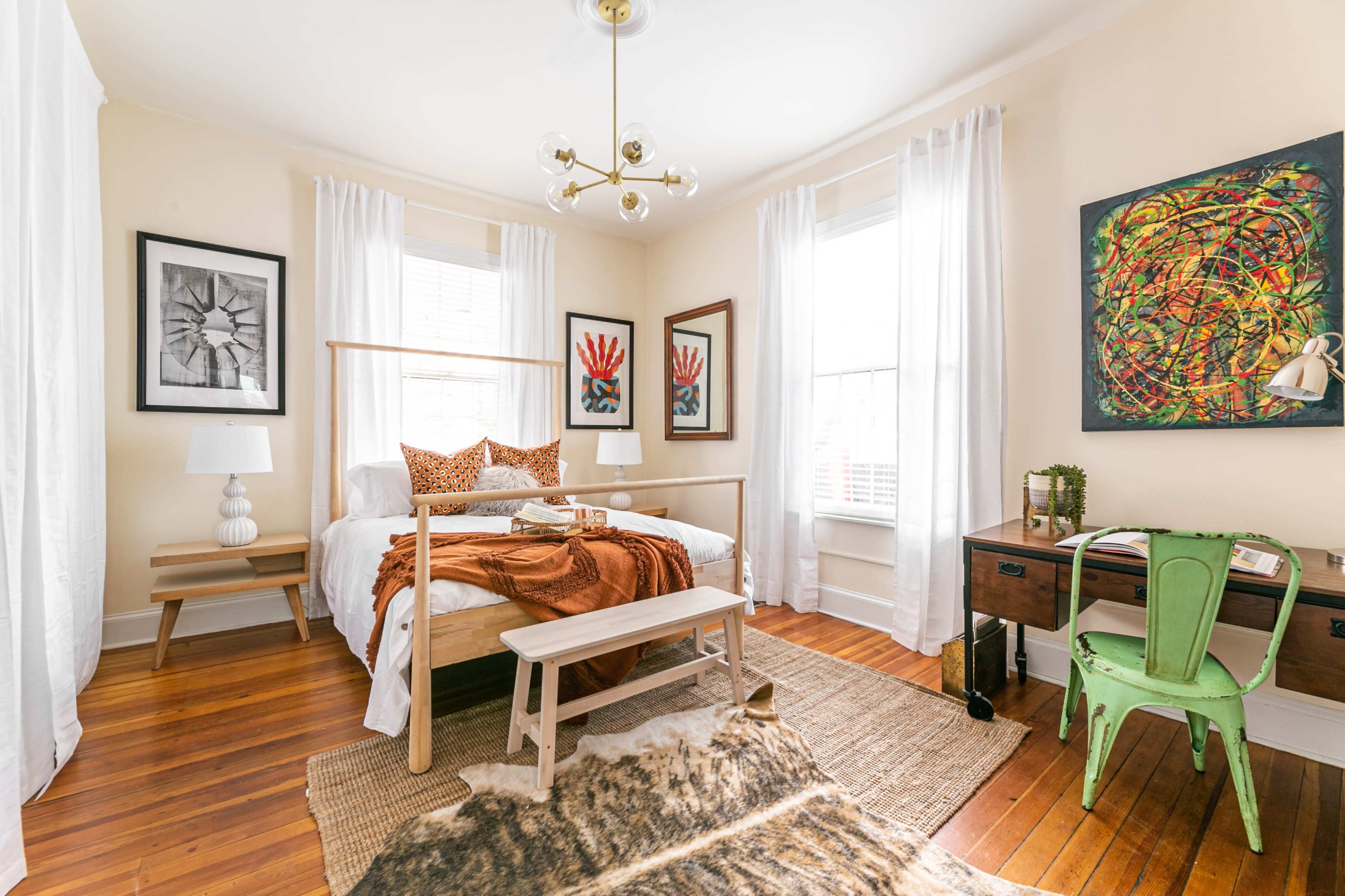 The room features a bed with a wooden frame, a desk, and decorative art on the walls, complemented by natural light from large windows.