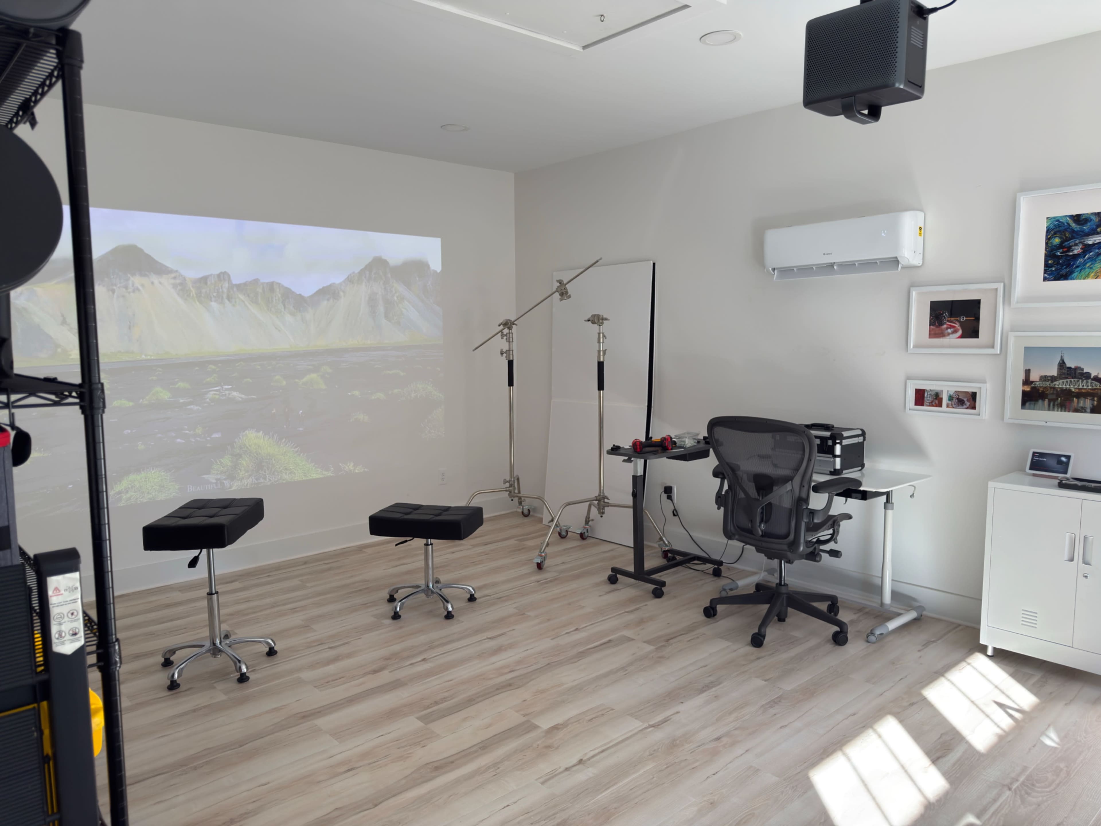 The room features two black stools, a desk with a chair, a backdrop stand, and a projected landscape image on the wall.