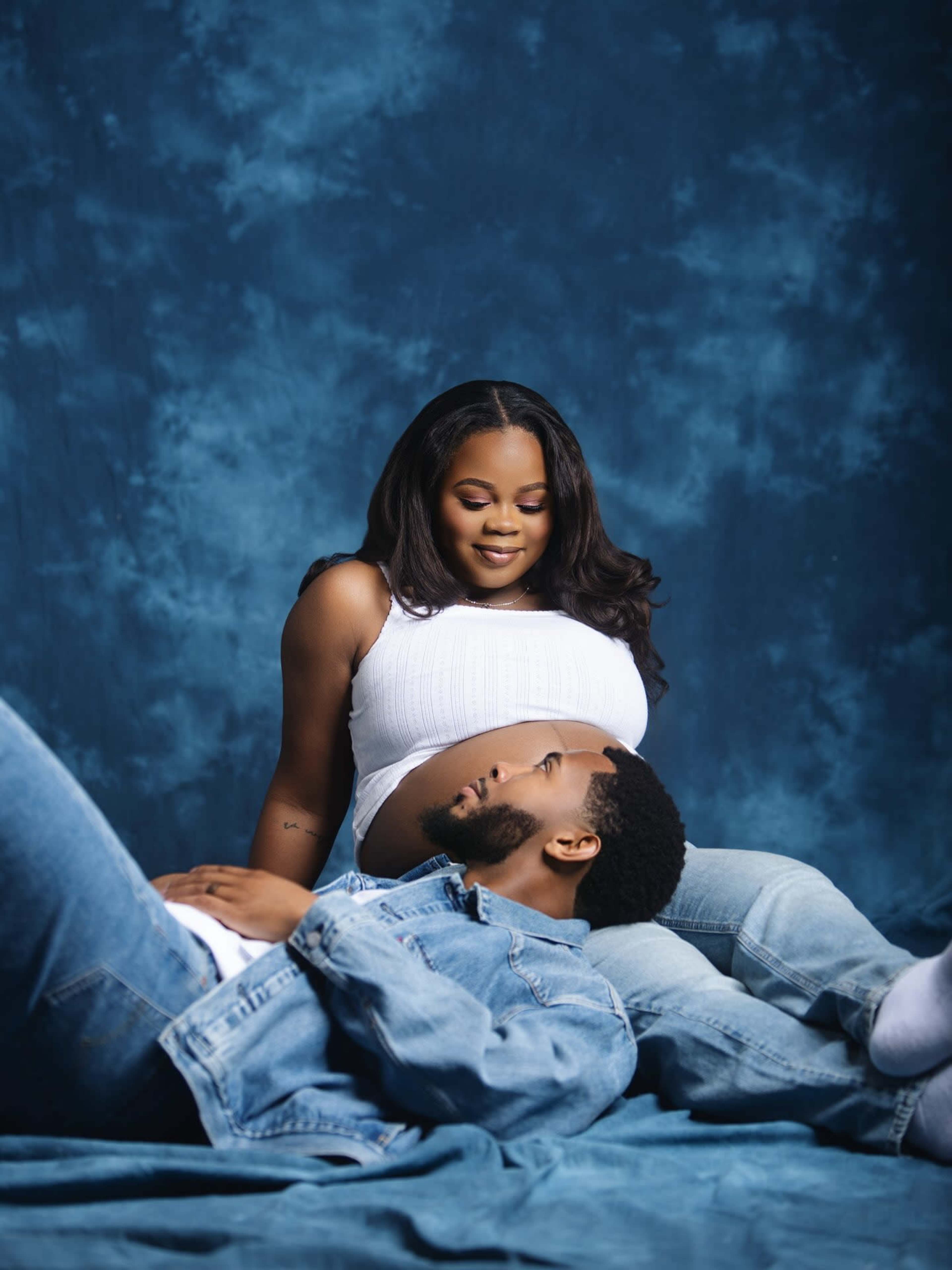A pregnant woman in a white top sits above a man in a denim jacket and blue jeans, both positioned on a blue backdrop.