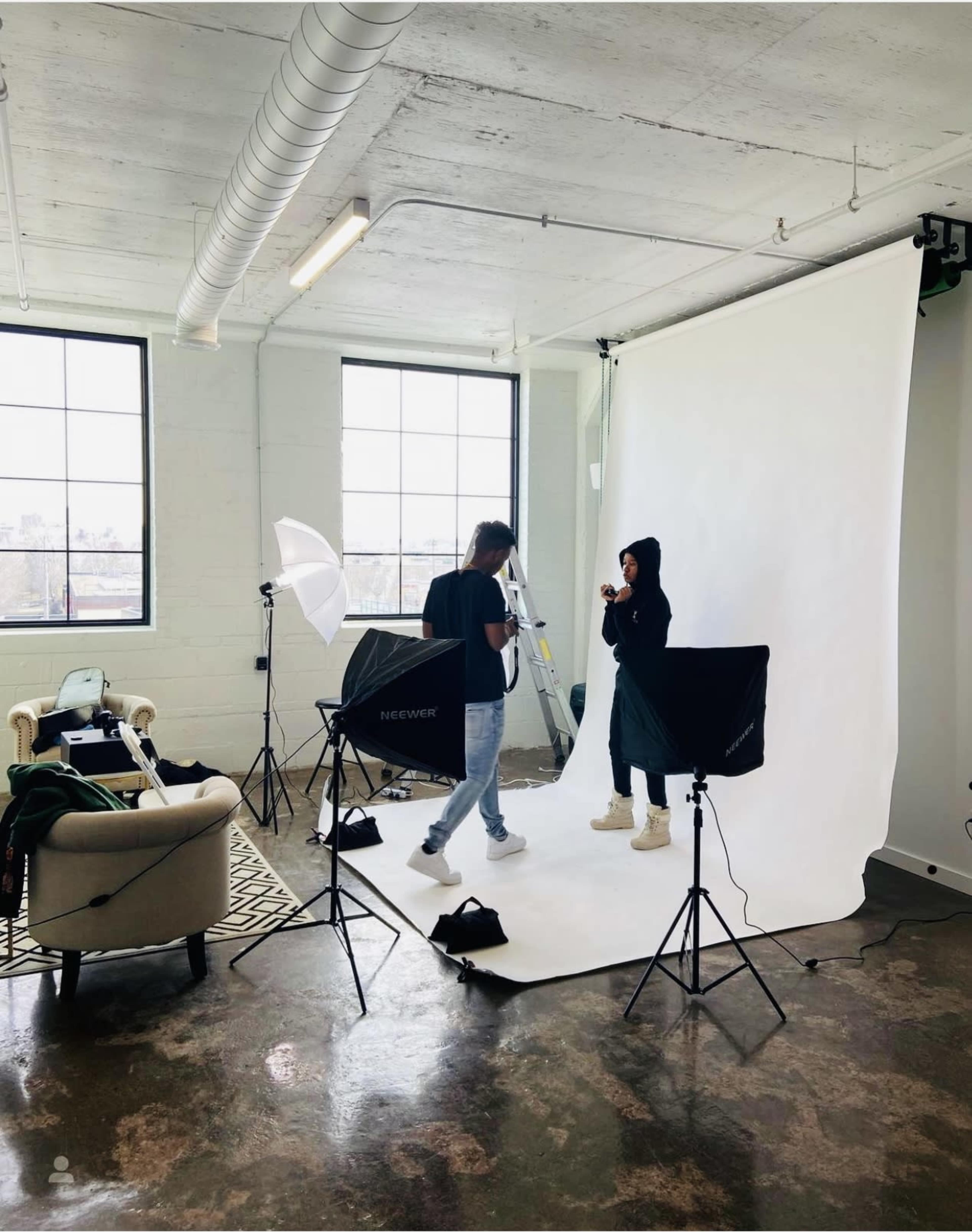A photography studio features a model and photographer preparing for a shoot with lighting equipment and a backdrop.