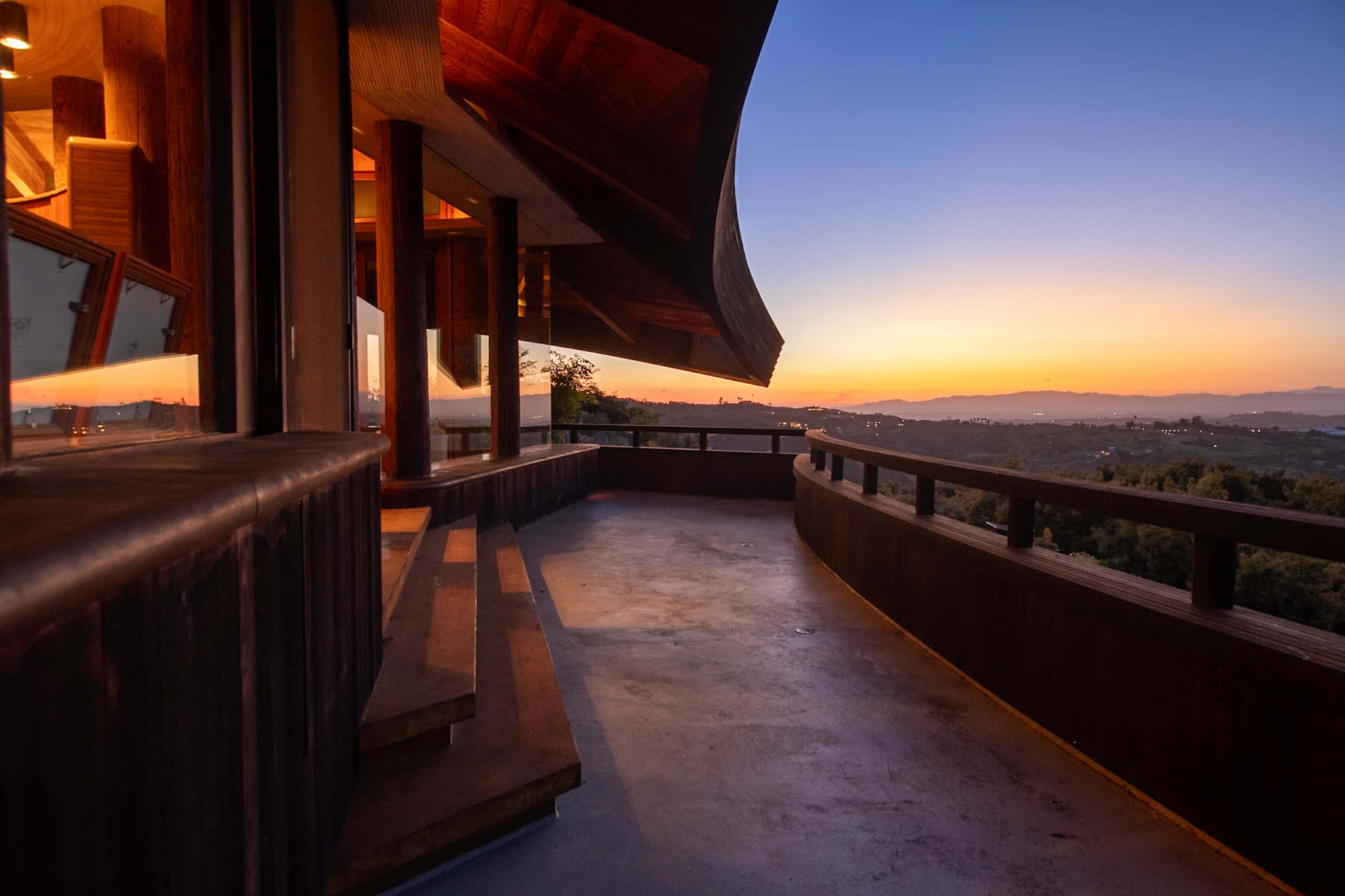 A wooden deck with curved railings overlooks a scenic landscape at sunset, highlighting the architectural features of the house.