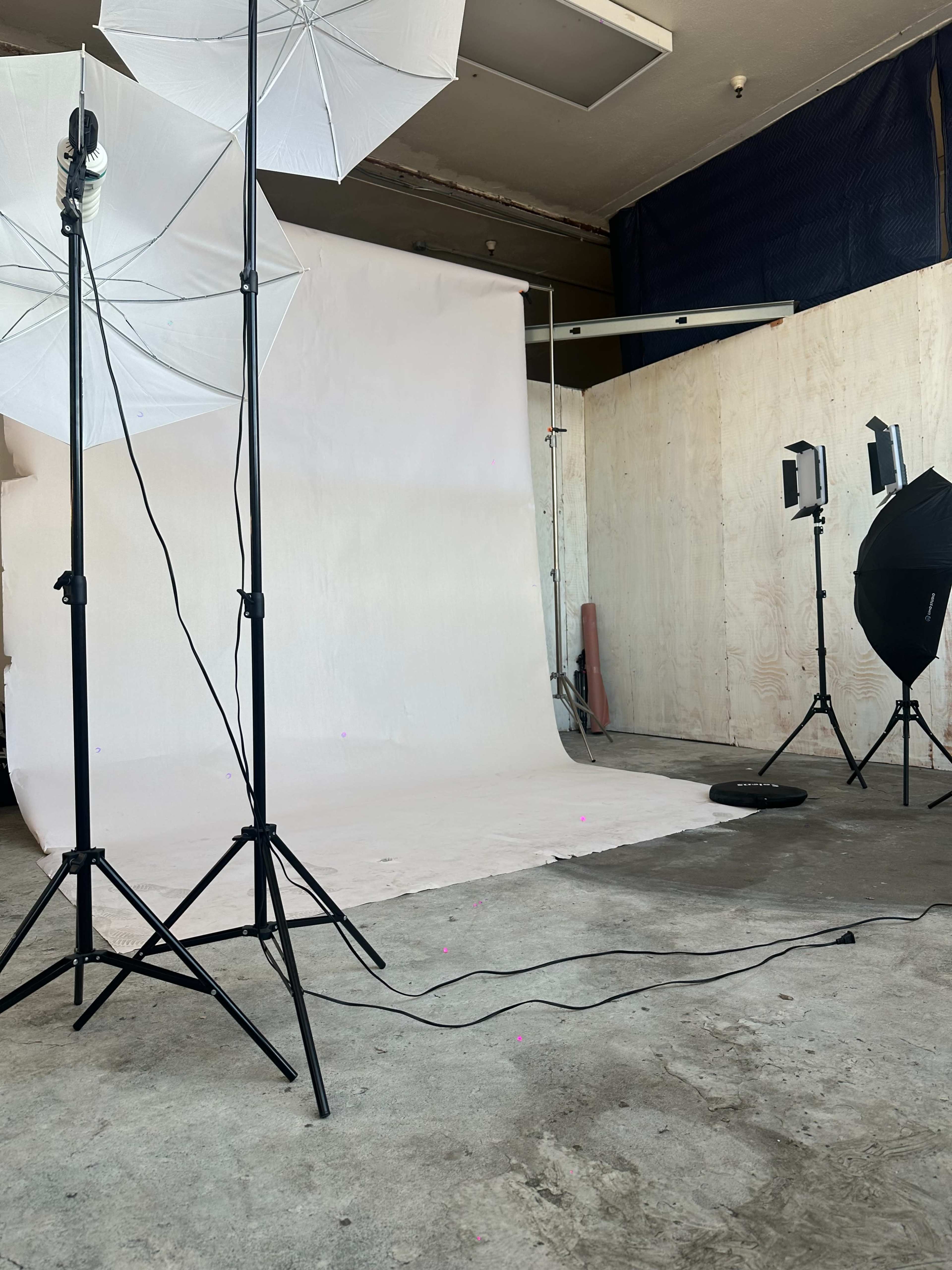 The image shows a photography studio with a white backdrop, lighting equipment, and cables on a concrete floor.
