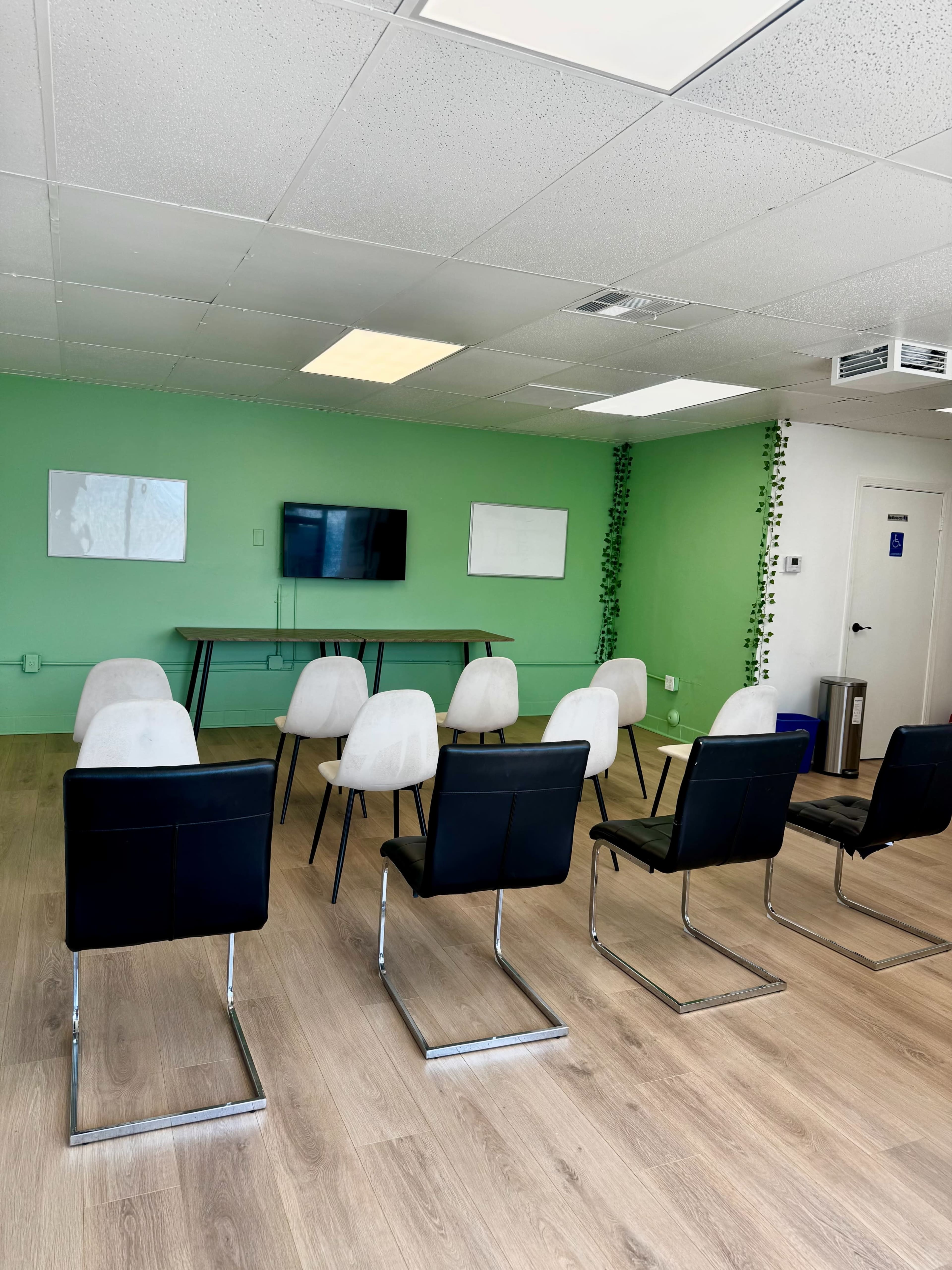 The image shows a meeting room with a green wall, a large table at the center, and rows of chairs arranged for seating.