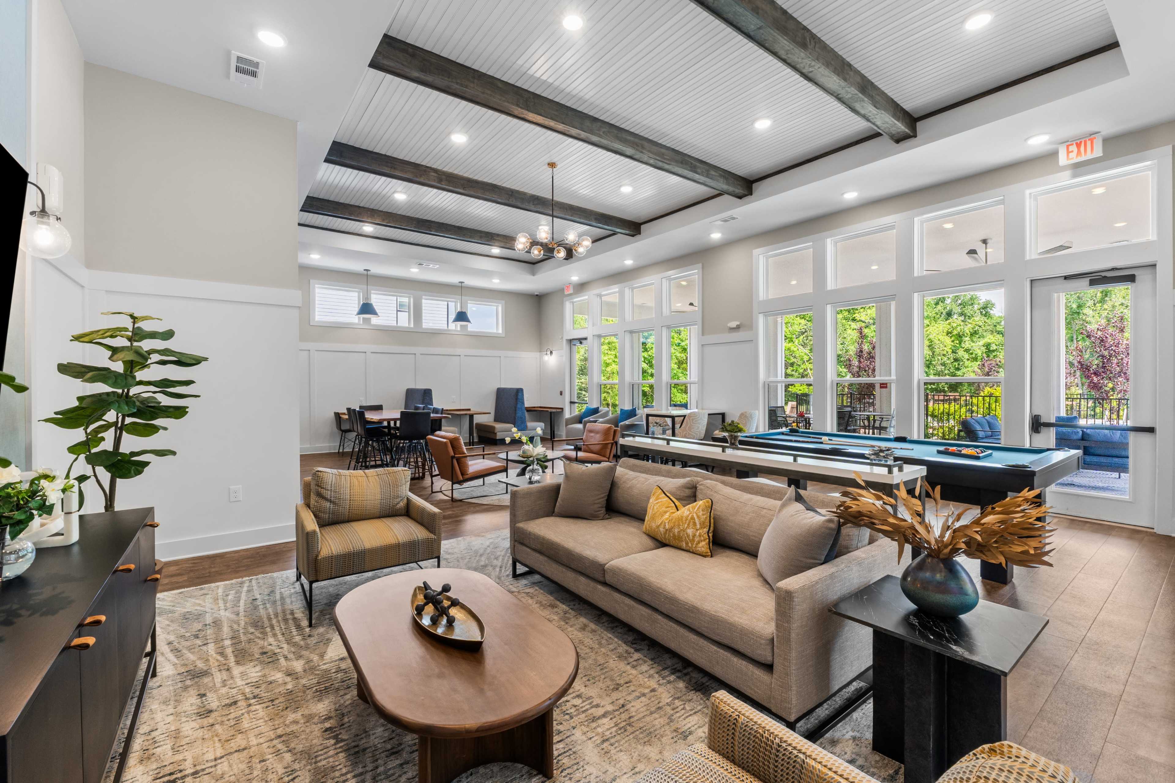 The image shows a spacious community lounge area featuring modern furniture, large windows, and an open layout with recreational tables.