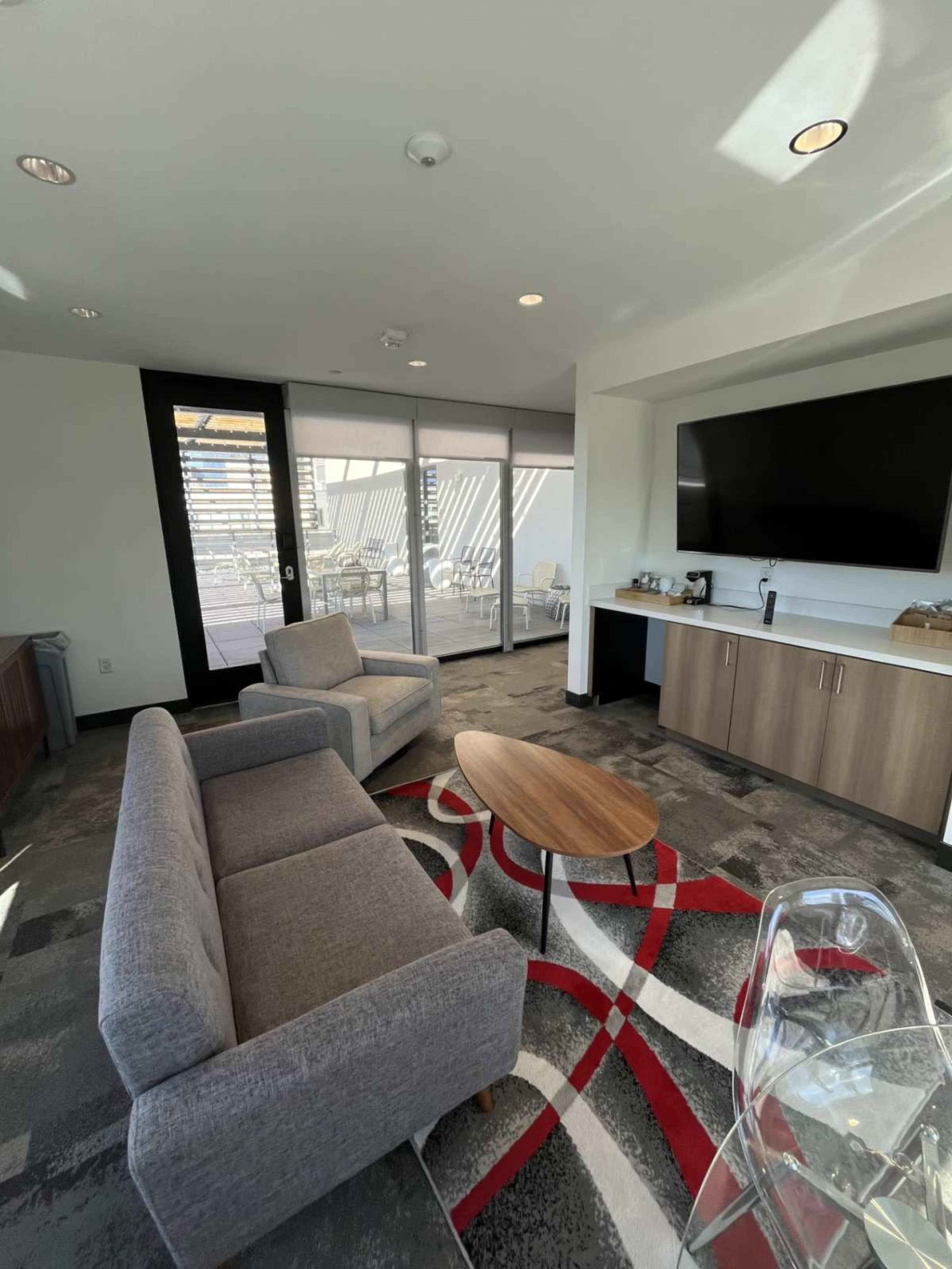 The image shows a modern lounge area with two gray sofas, a wooden coffee table, a large television mounted on the wall, and a glass door leading to an outdoor patio.