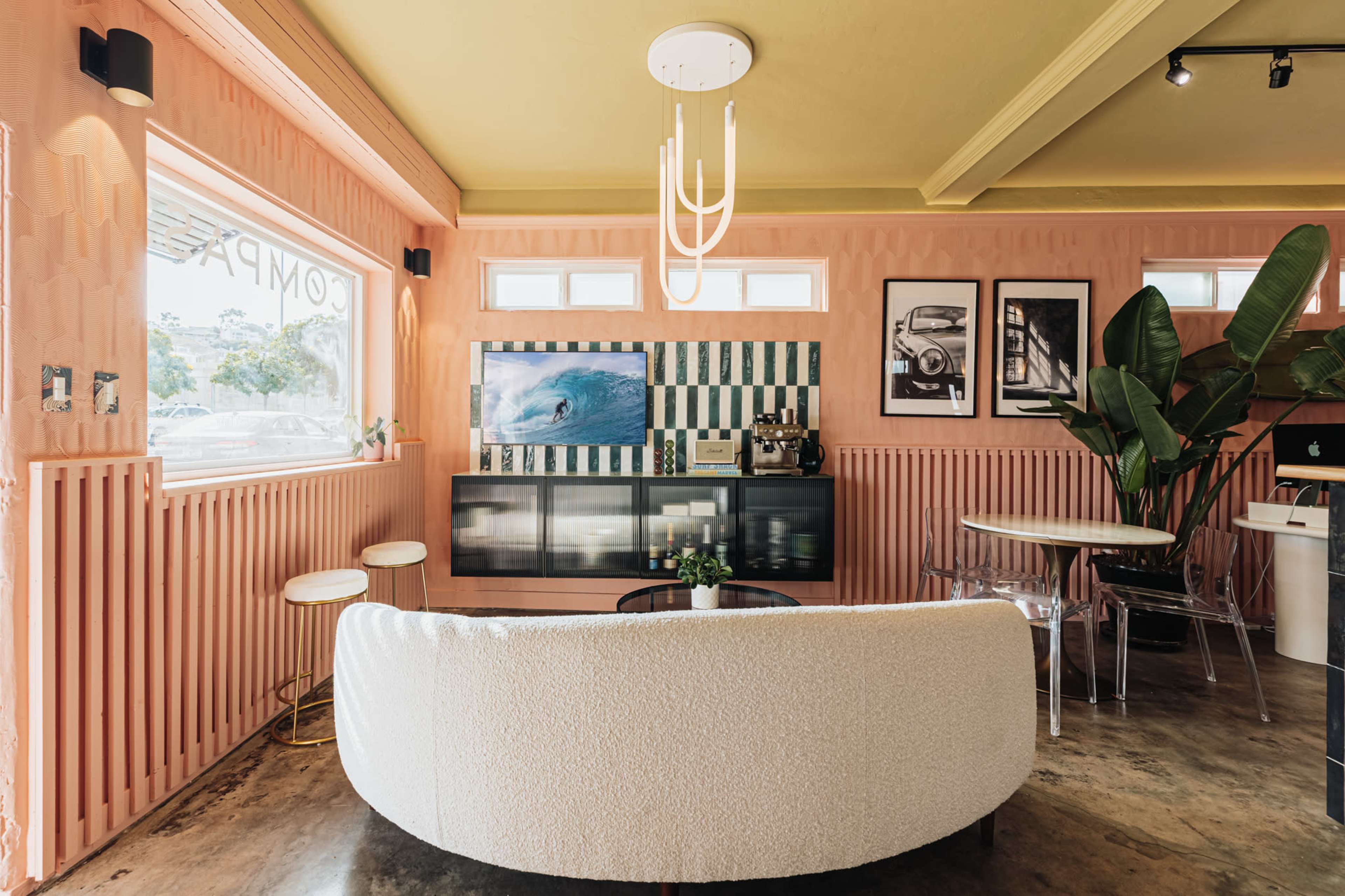 The image shows a modern interior space with a curved white sofa, a coffee station, and large windows, all accented by textured pink walls and greenery.