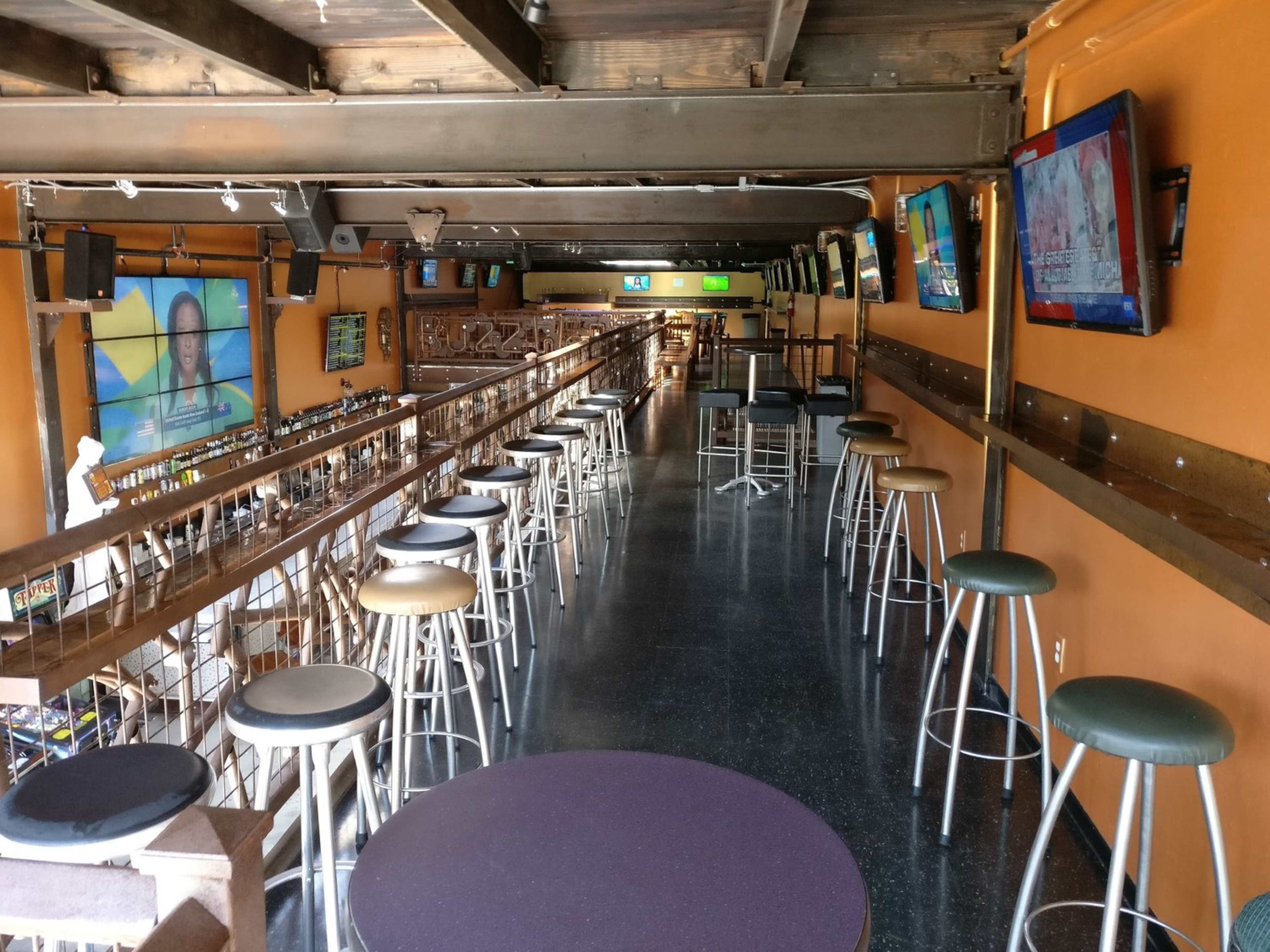 The image shows an empty bar interior with high stools lining a long counter and several televisions mounted on the walls.