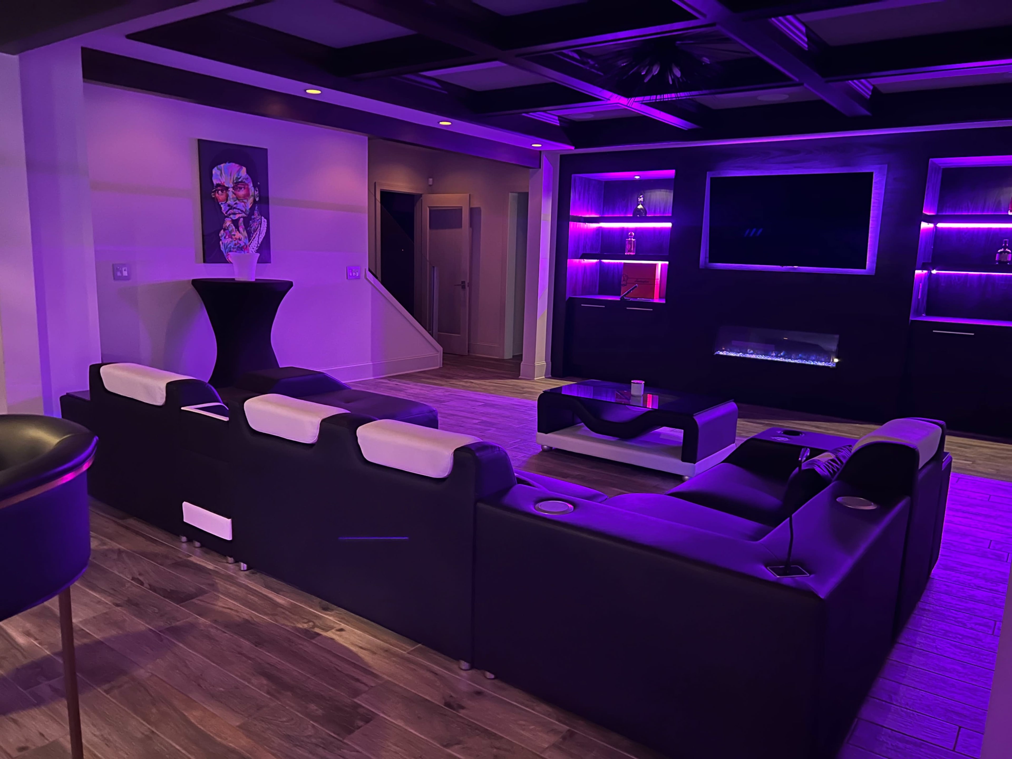 A modern living room with a black sectional sofa, a flat-screen television mounted on the wall, and purple ambient lighting.