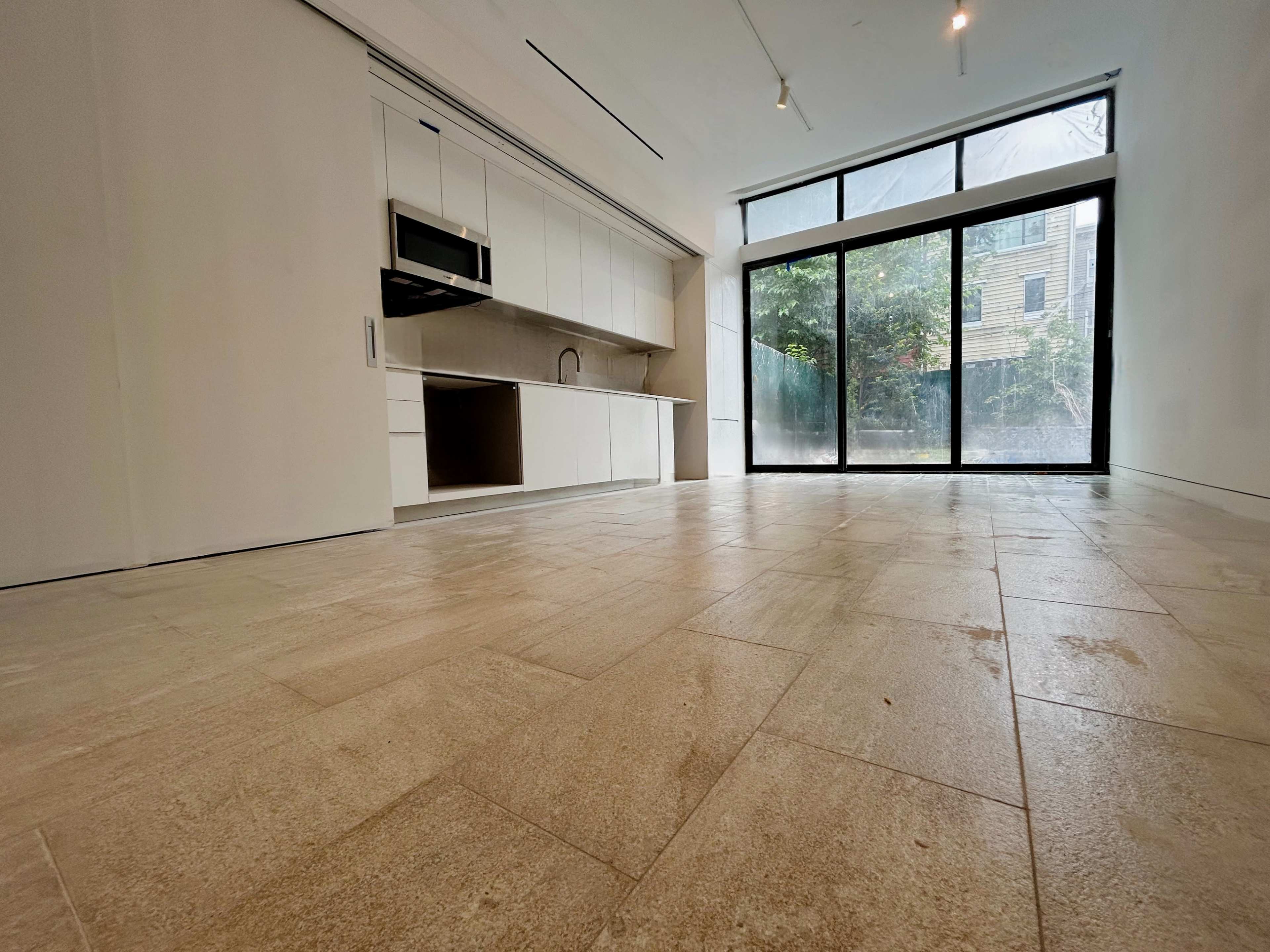 The image shows a modern, empty interior space with a kitchen area on one side and large sliding glass doors leading to an outdoor view.
