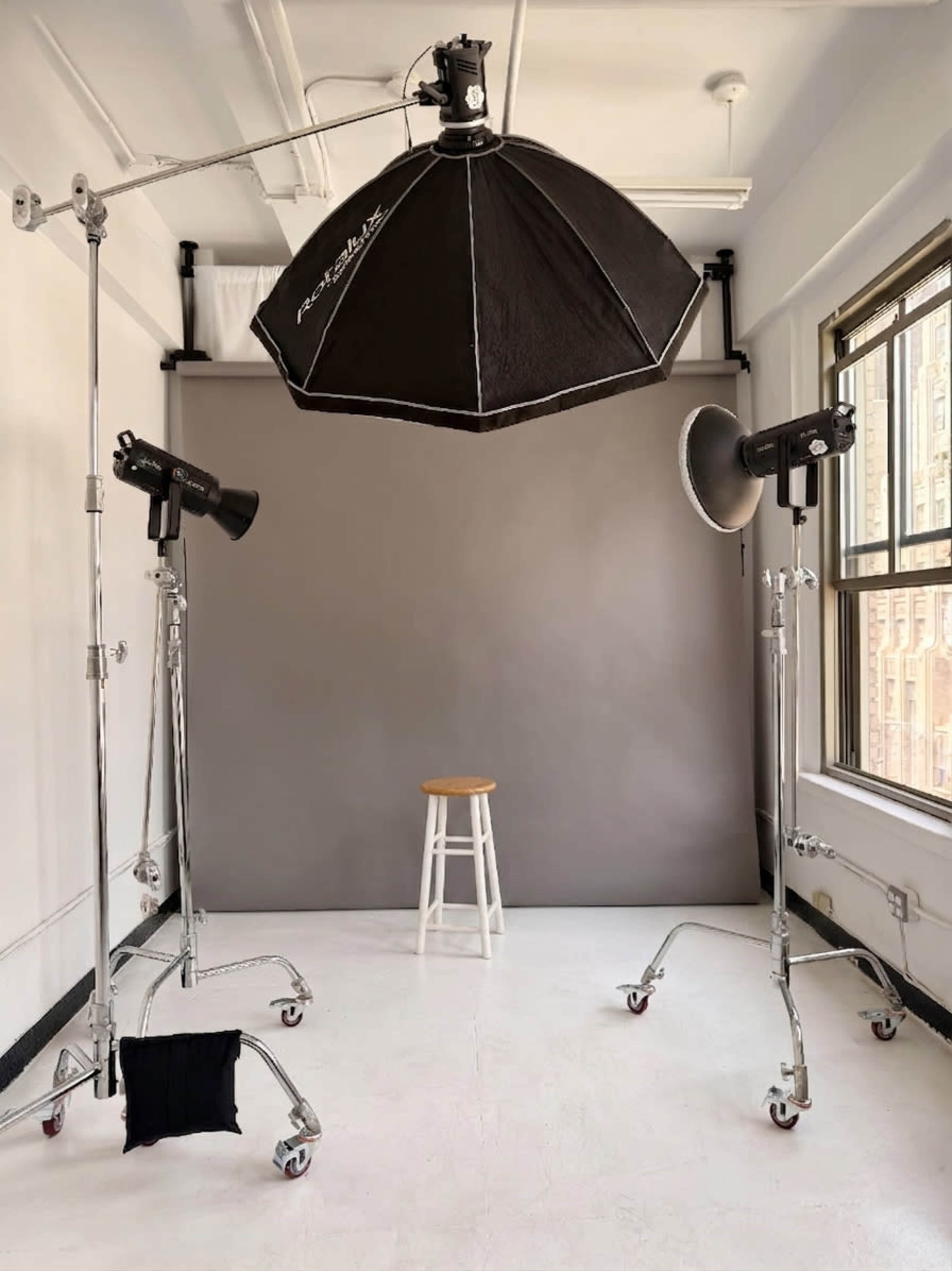 The image shows a photography studio setup featuring a gray backdrop, a wooden stool, and multiple light sources mounted on stands.
