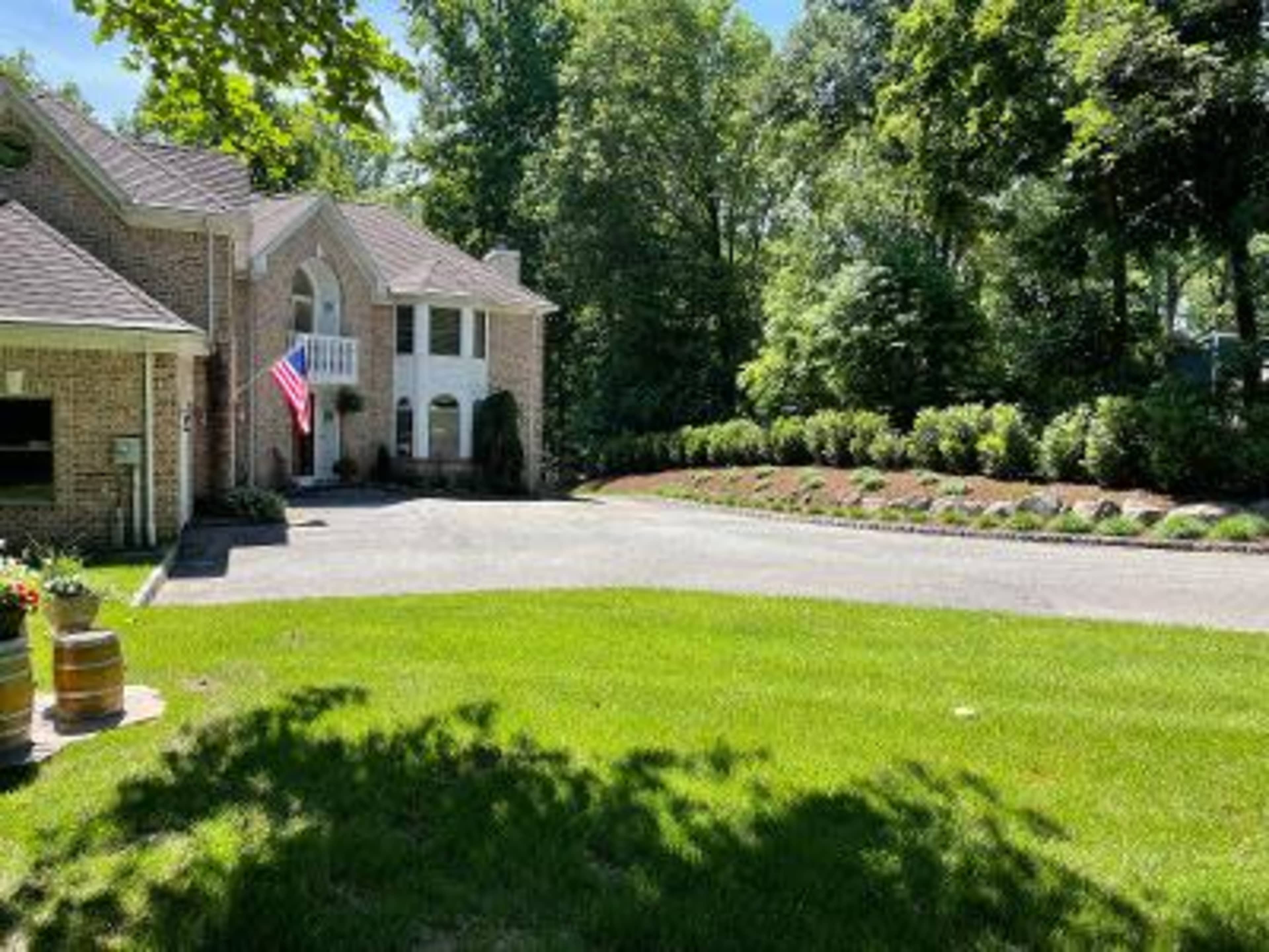 A brick house with a front porch and an American flag displays a well-maintained driveway and a landscaped garden lined with shrubs.