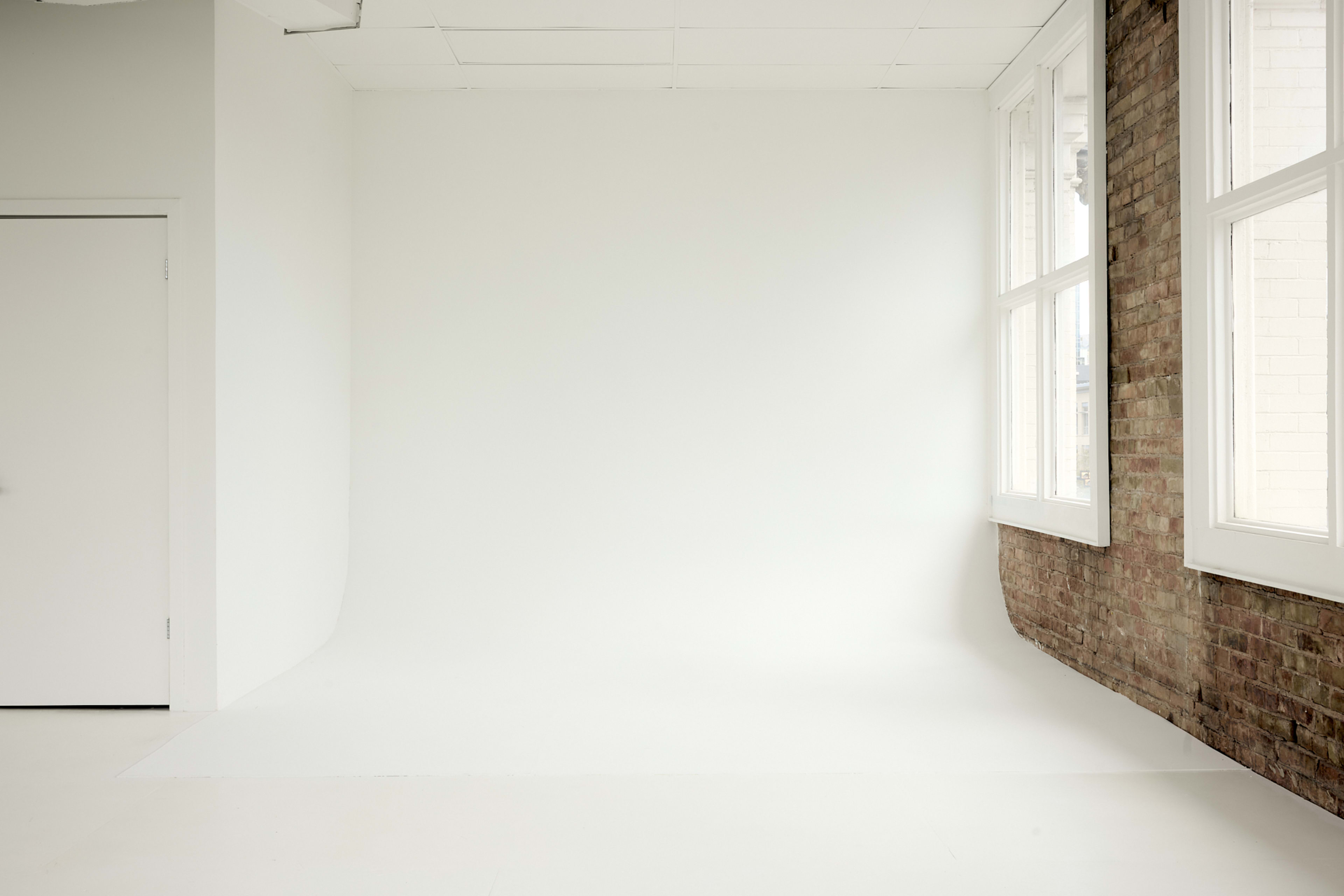 The image shows a photography studio with a white backdrop and large windows on one side, complemented by a brick wall.