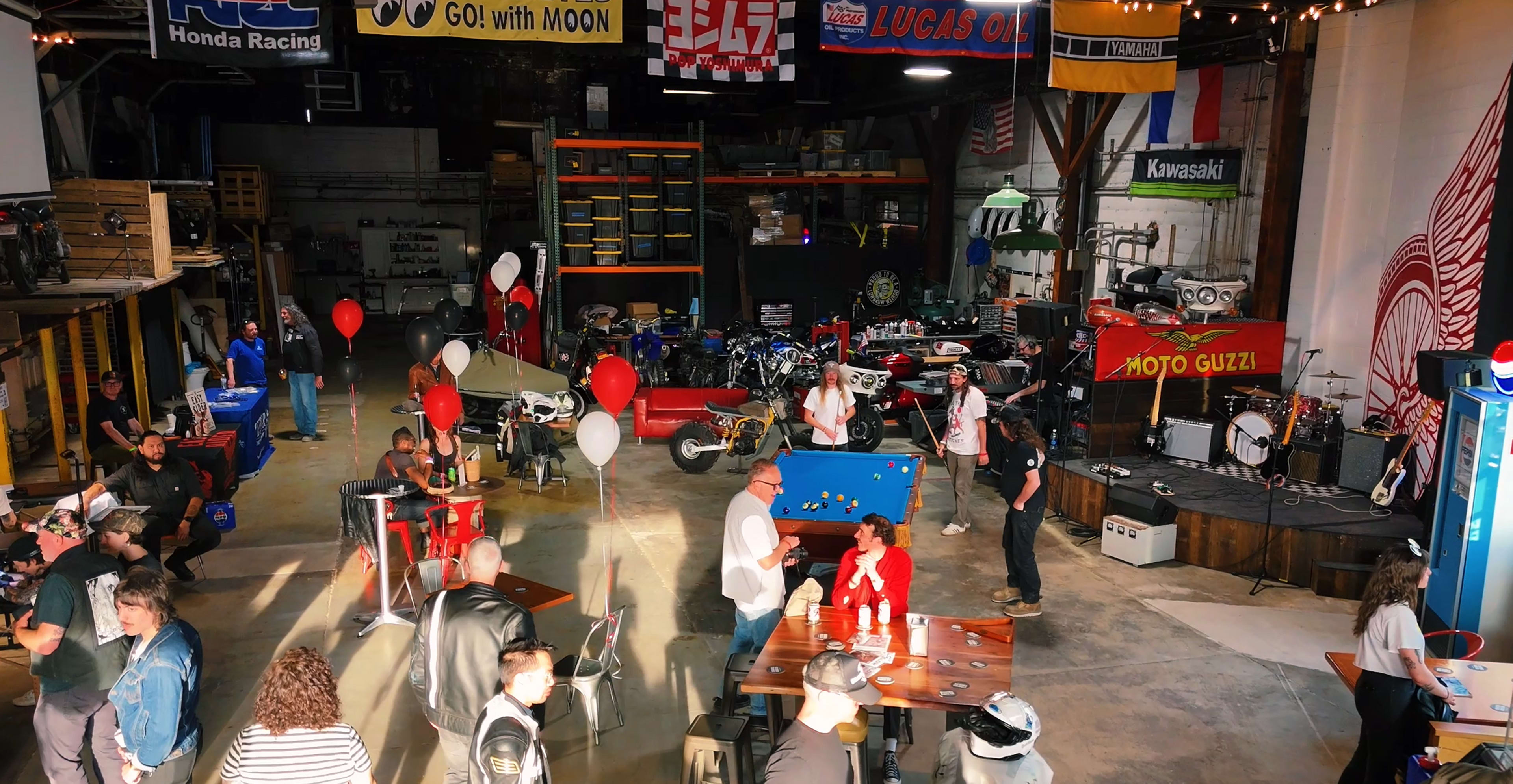 A large garage space is filled with people socializing, playing games, and surrounded by various motorcycles and banners.