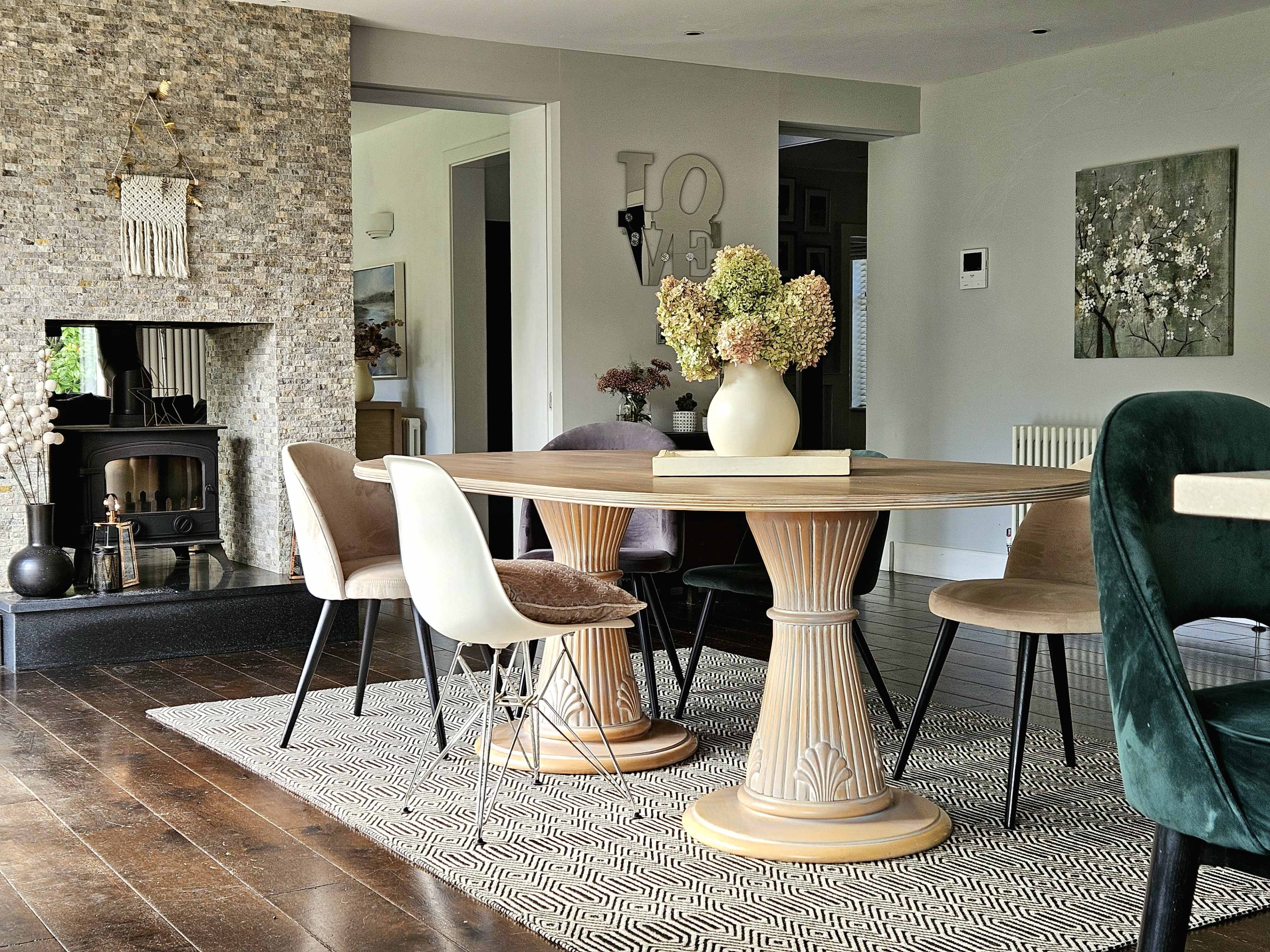 The image shows a modern dining area with a round table surrounded by chairs, a patterned rug, and a stone fireplace in the background.