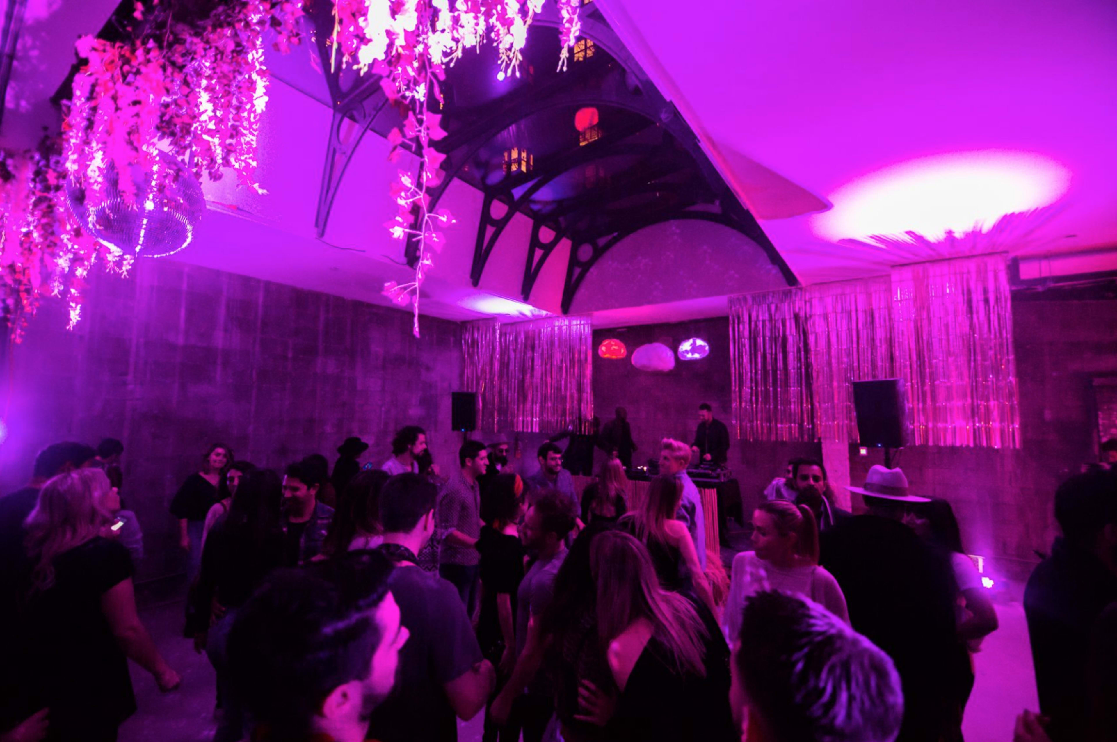 A crowded indoor venue is illuminated by purple lighting, featuring a DJ setup and decorative elements like hanging flowers and glittering streamers.
