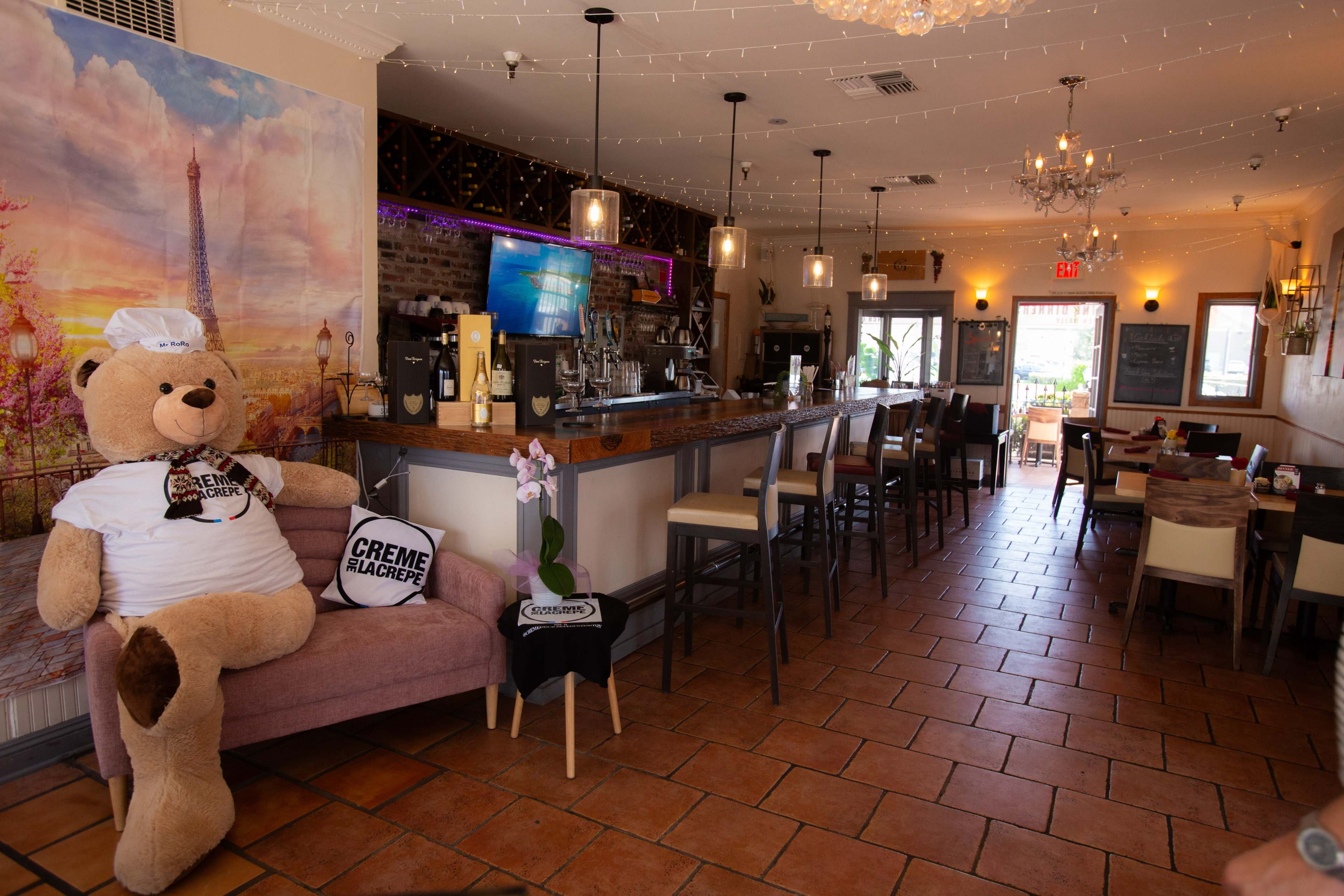 The image shows the interior of a café featuring a large teddy bear dressed as a chef sitting on a couch beside a bar area, with tables and chairs arranged in the background.