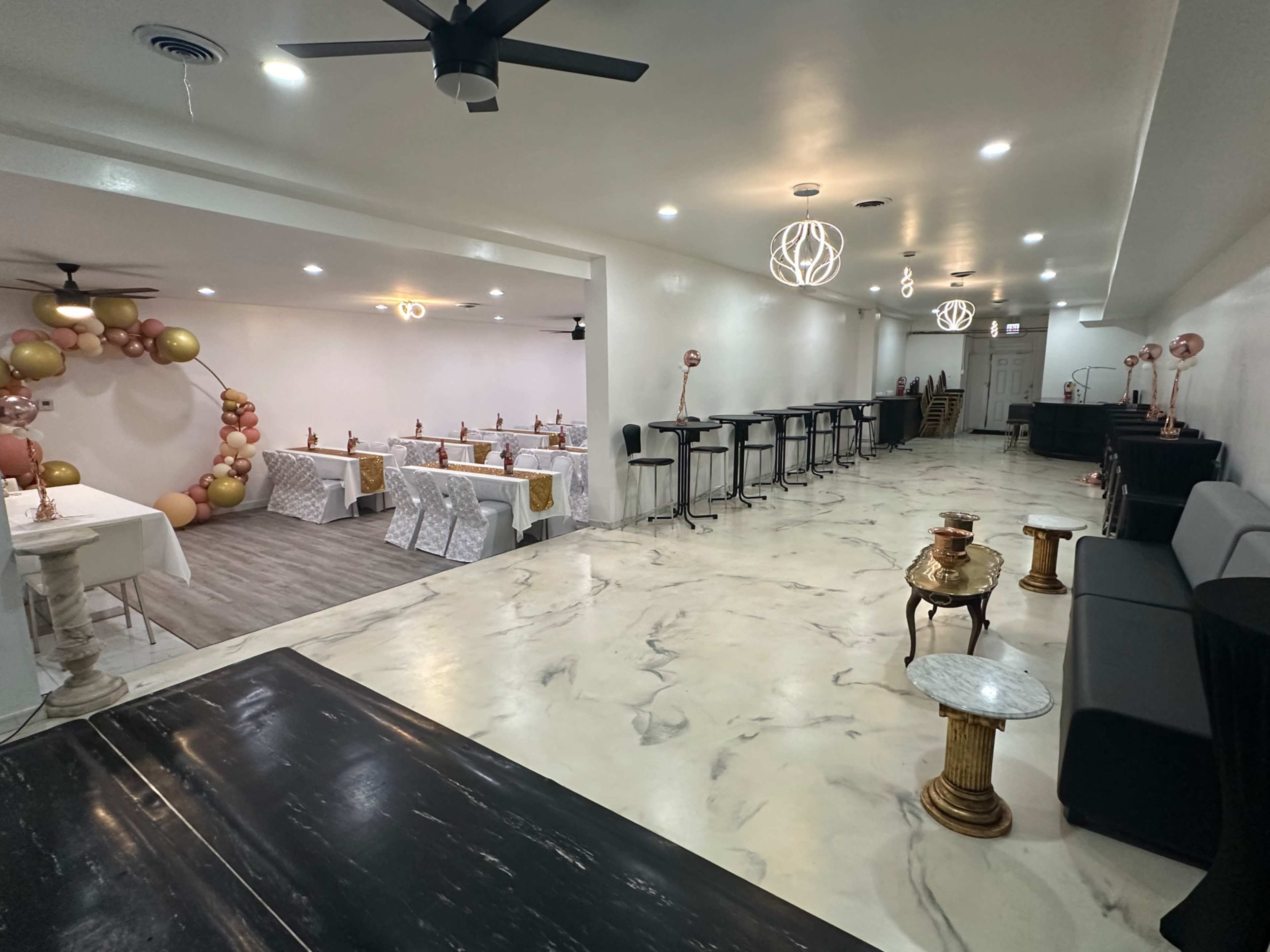 The image shows an elegantly designed event space featuring tables with white tablecloths, decorative balloons, and seating areas arranged around the room.