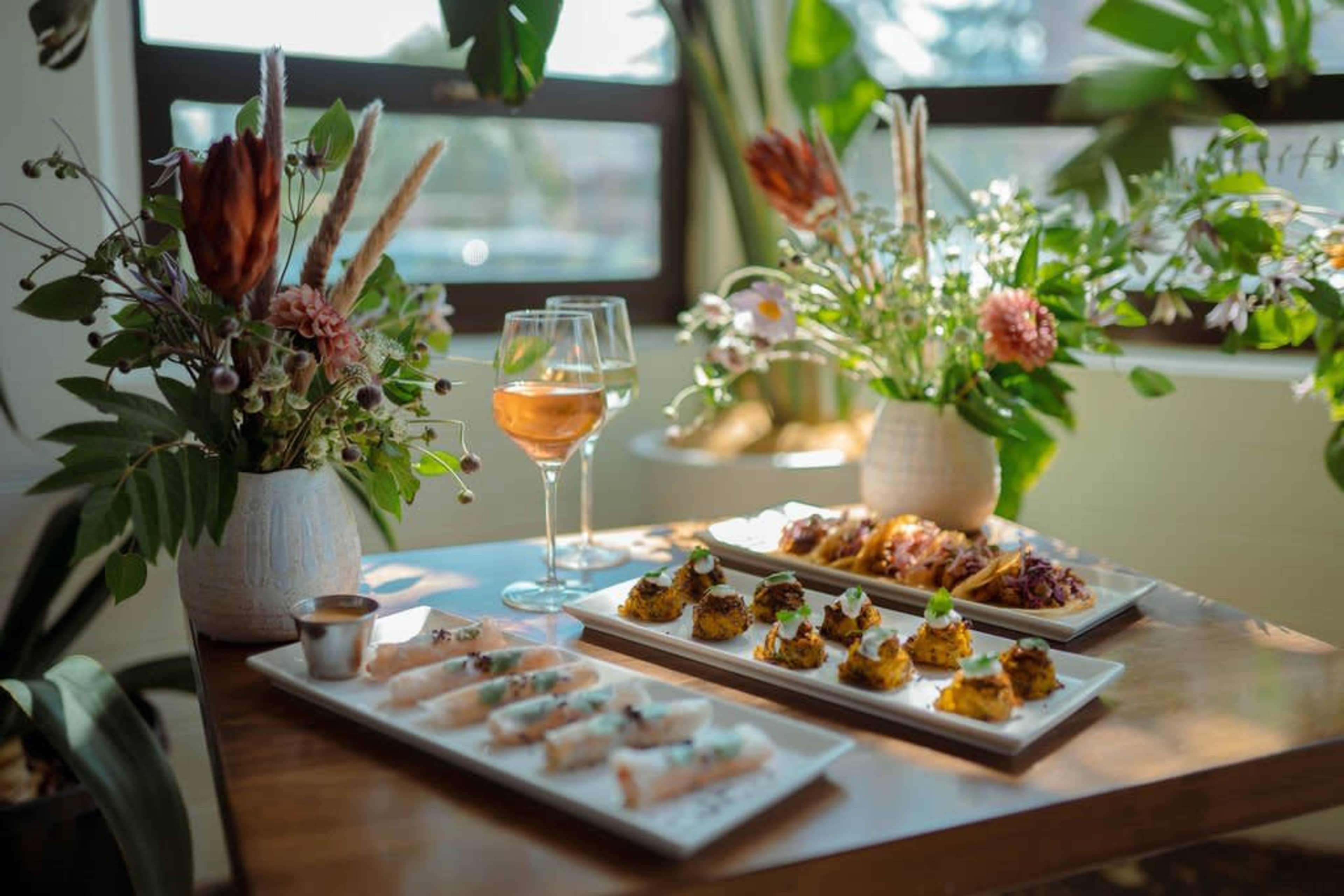 A wooden table is set with several plates of appetizers and two glasses of wine, surrounded by decorative plants and floral arrangements.