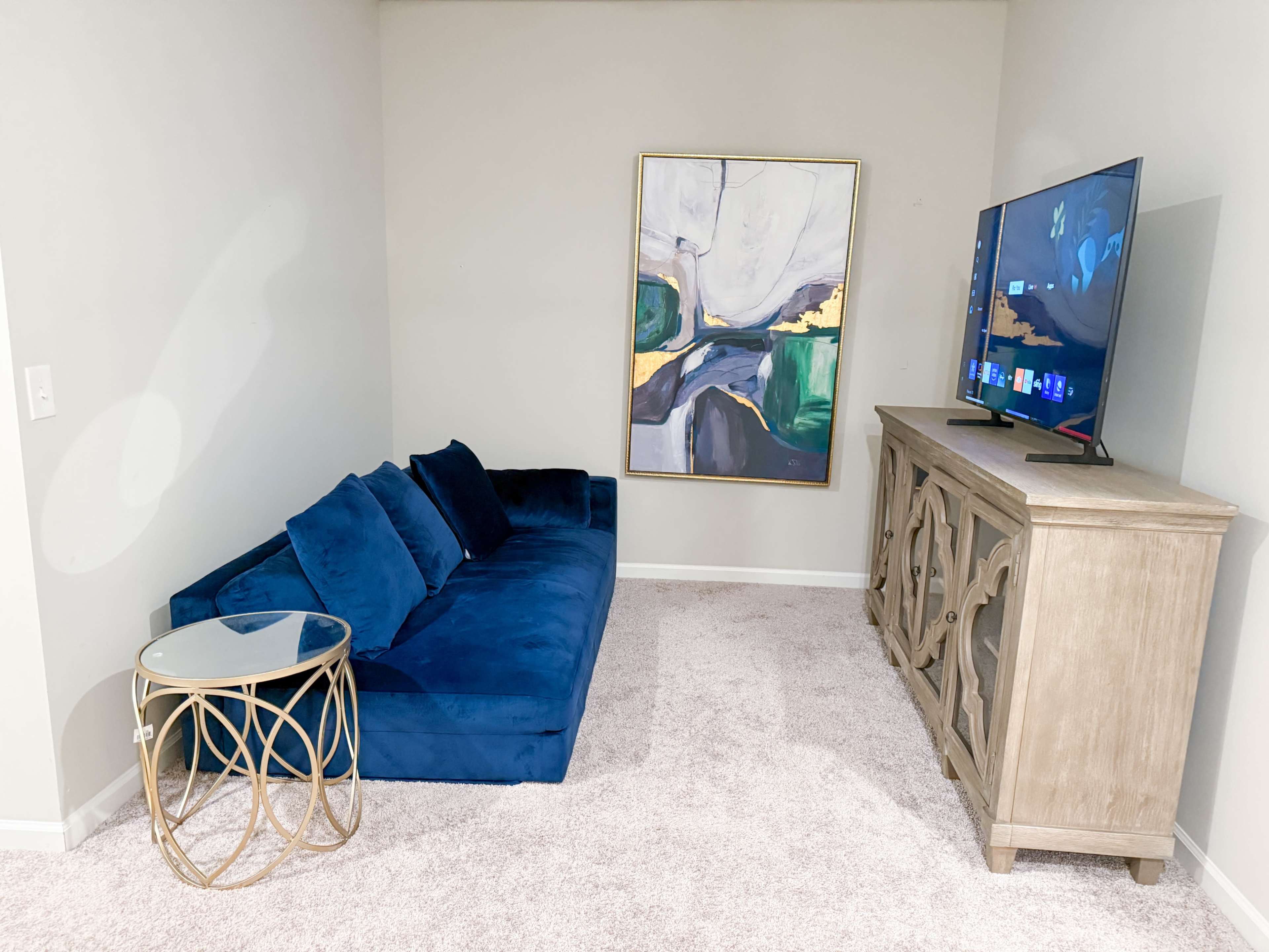 The image shows a small living space with a blue velvet sofa, a glass side table, a decorative wooden cabinet, and a wall-mounted television alongside an abstract painting.