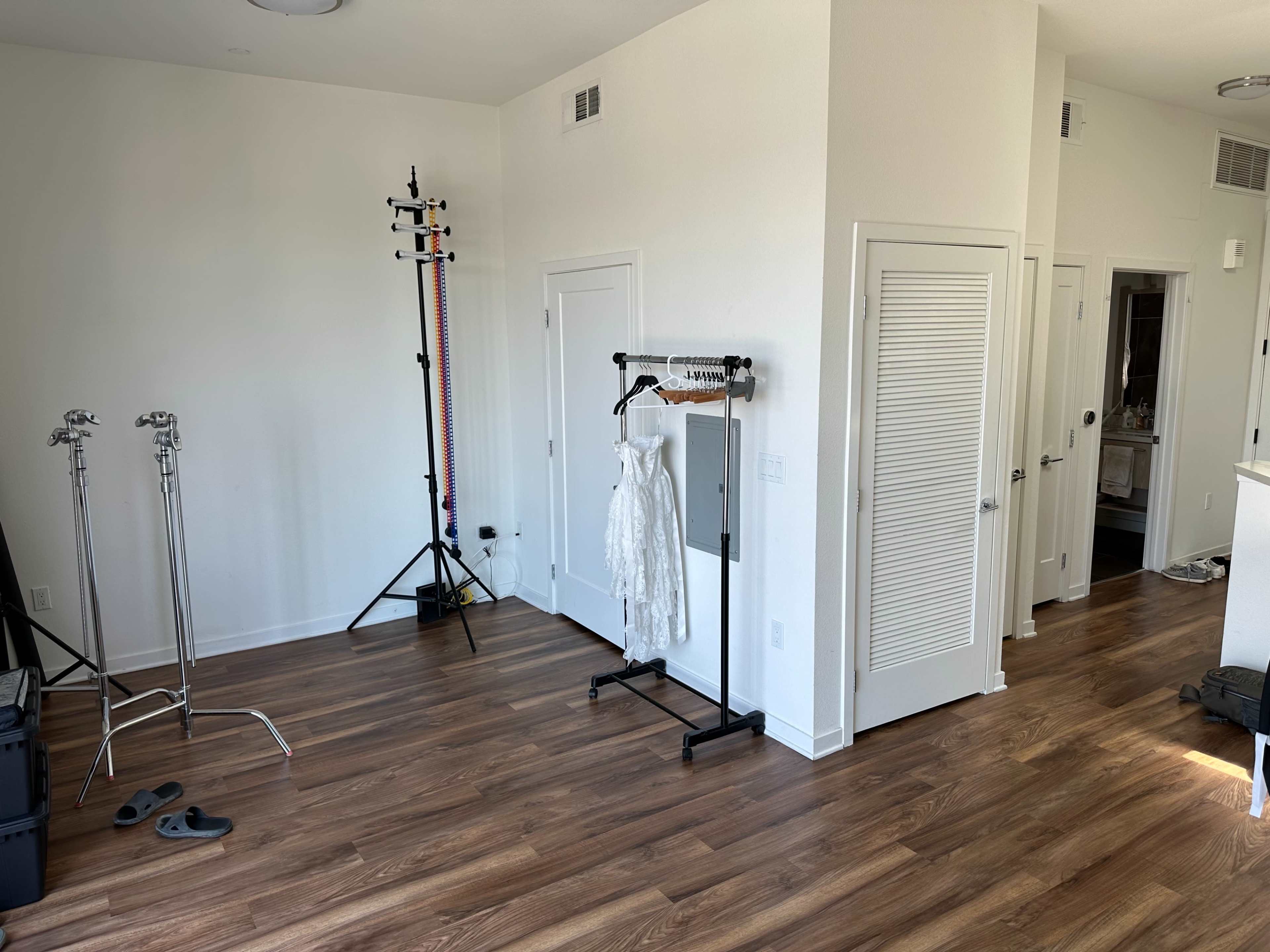 The image shows a well-lit room with wooden flooring, featuring a dress on a rack, two lighting stands, and doorways leading to other areas.