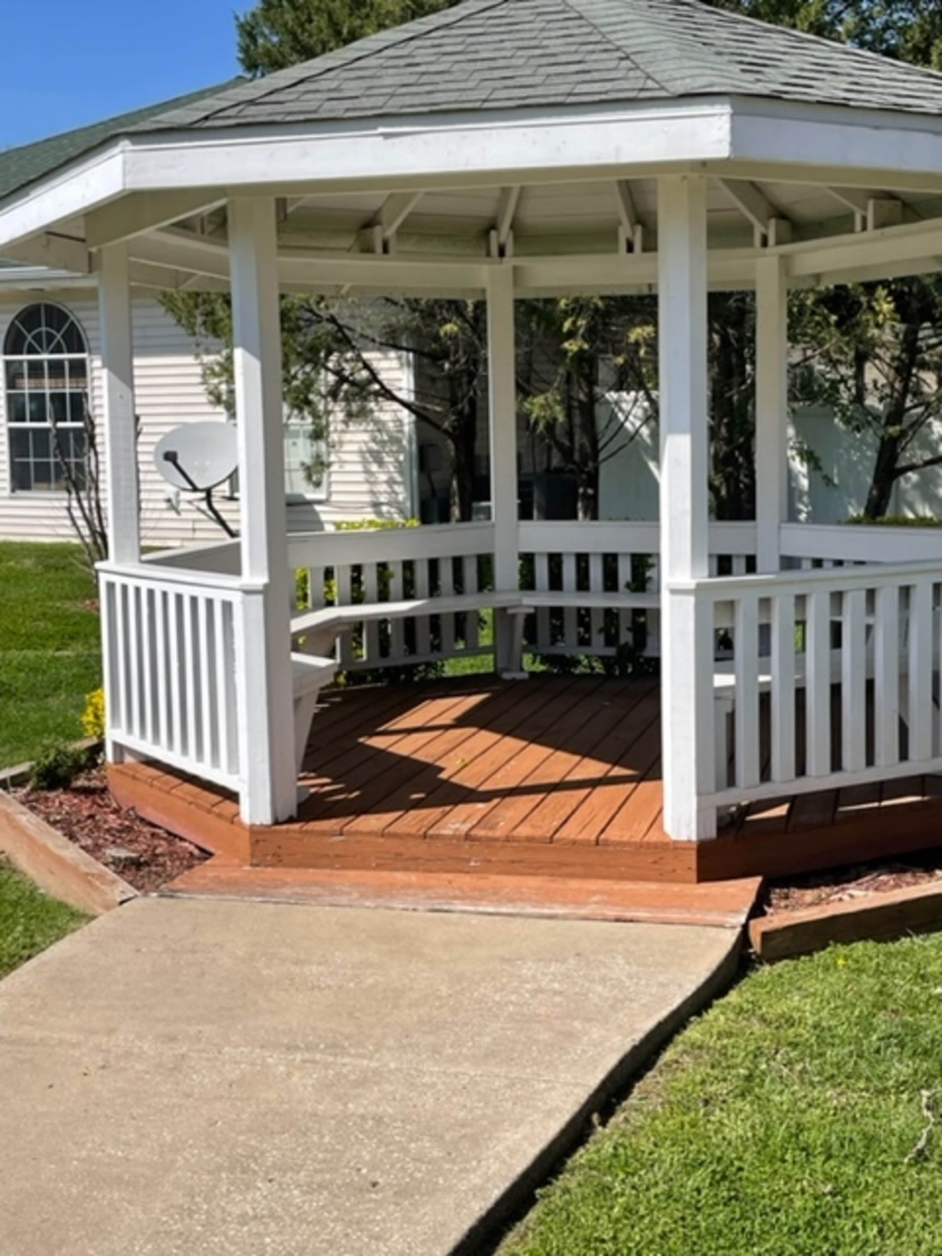A white wooden gazebo with a shingled roof sits on a concrete pathway surrounded by grass and garden beds.