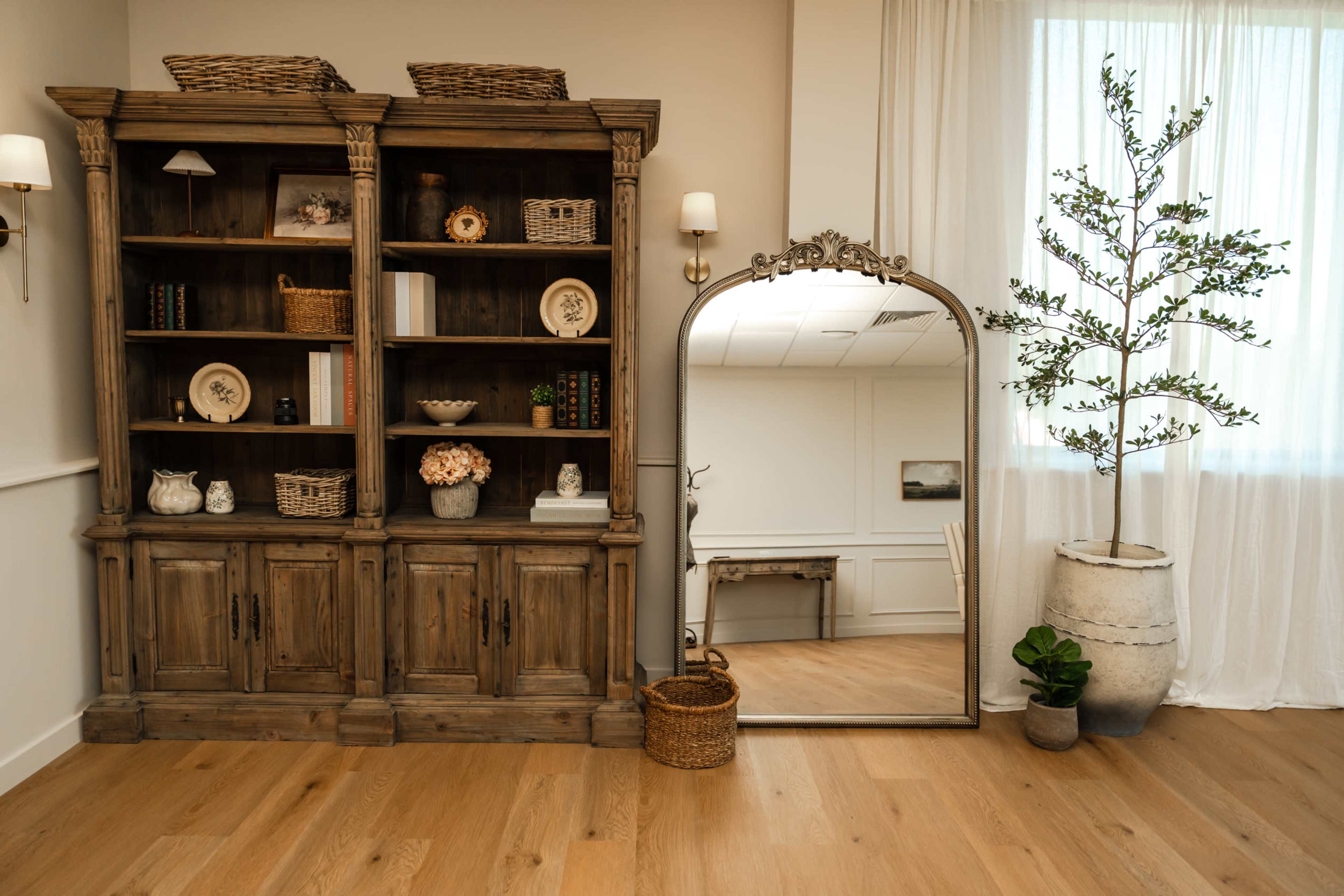 The image depicts a room featuring a wooden bookshelf with decorative items, a large ornate mirror, a potted plant, and a basket, all arranged against a wall with natural light coming through a window.