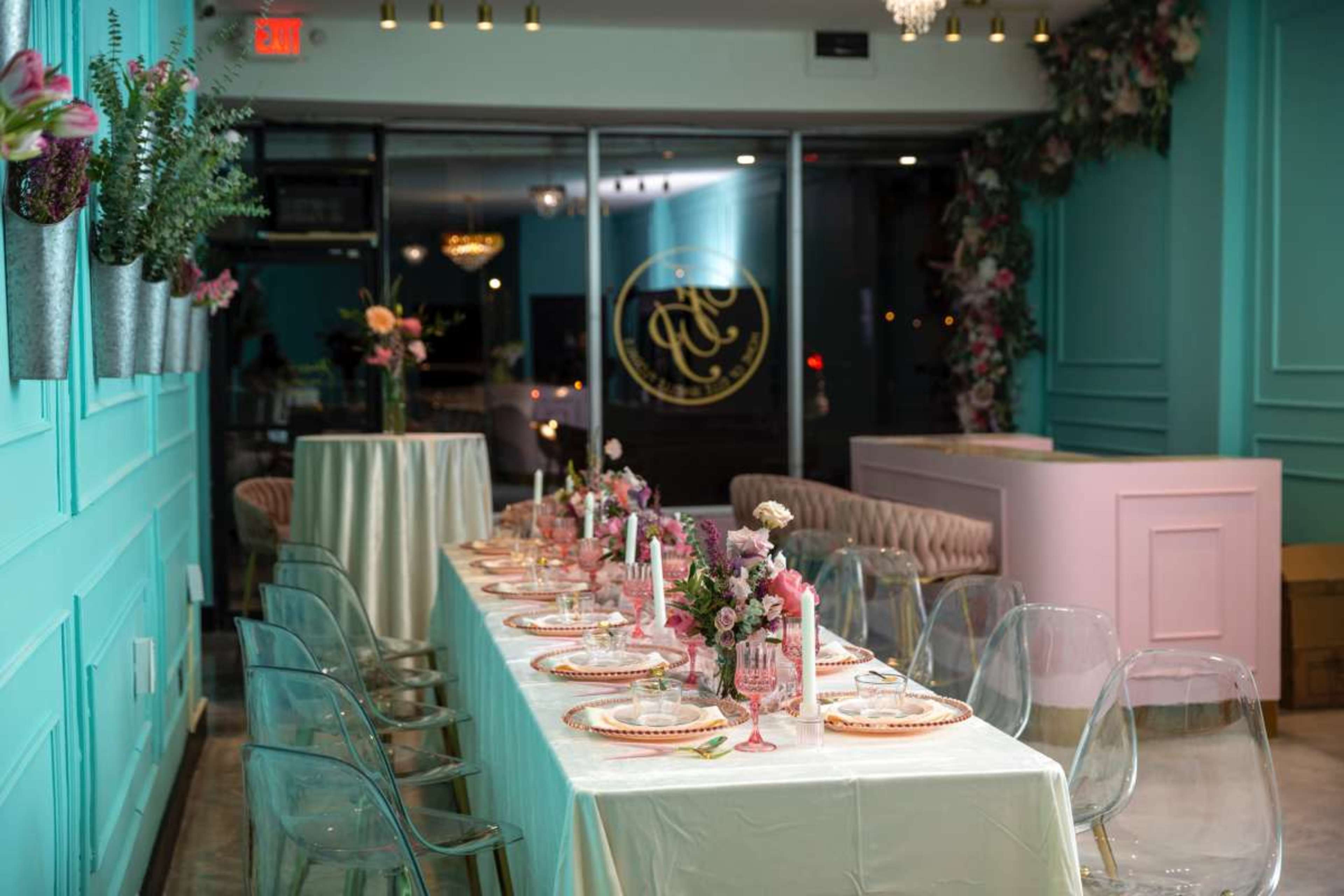 A long dining table is set with elegant dishware, candles, and flowers in a brightly colored boutique space with mint green walls and modern decor.