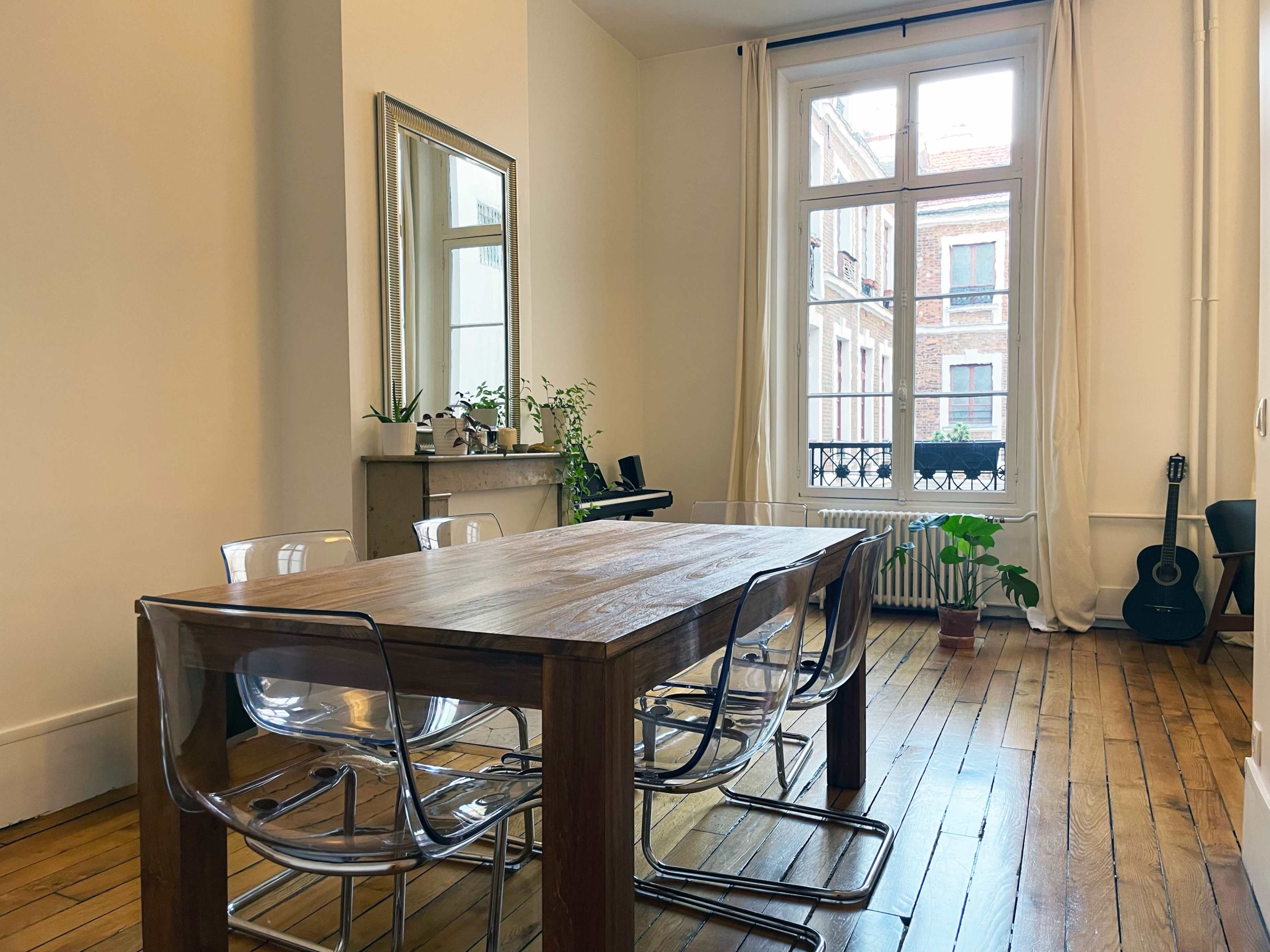 A wooden dining table with clear chairs is positioned in a room that features a large window overlooking a street, with plants and a guitar in the background.