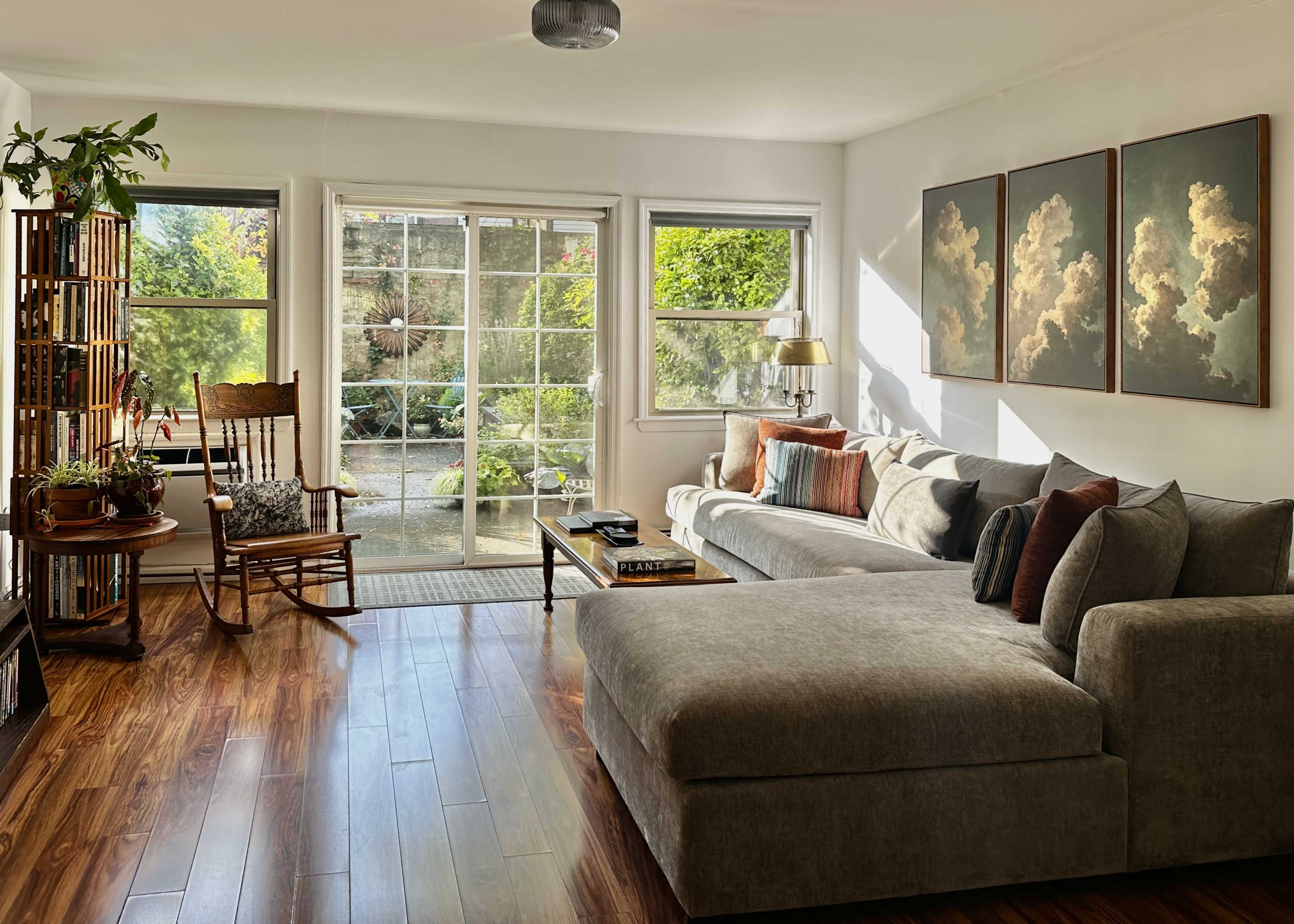 The image shows a bright living room with a sectional couch, a rocking chair, and large windows displaying a garden view.