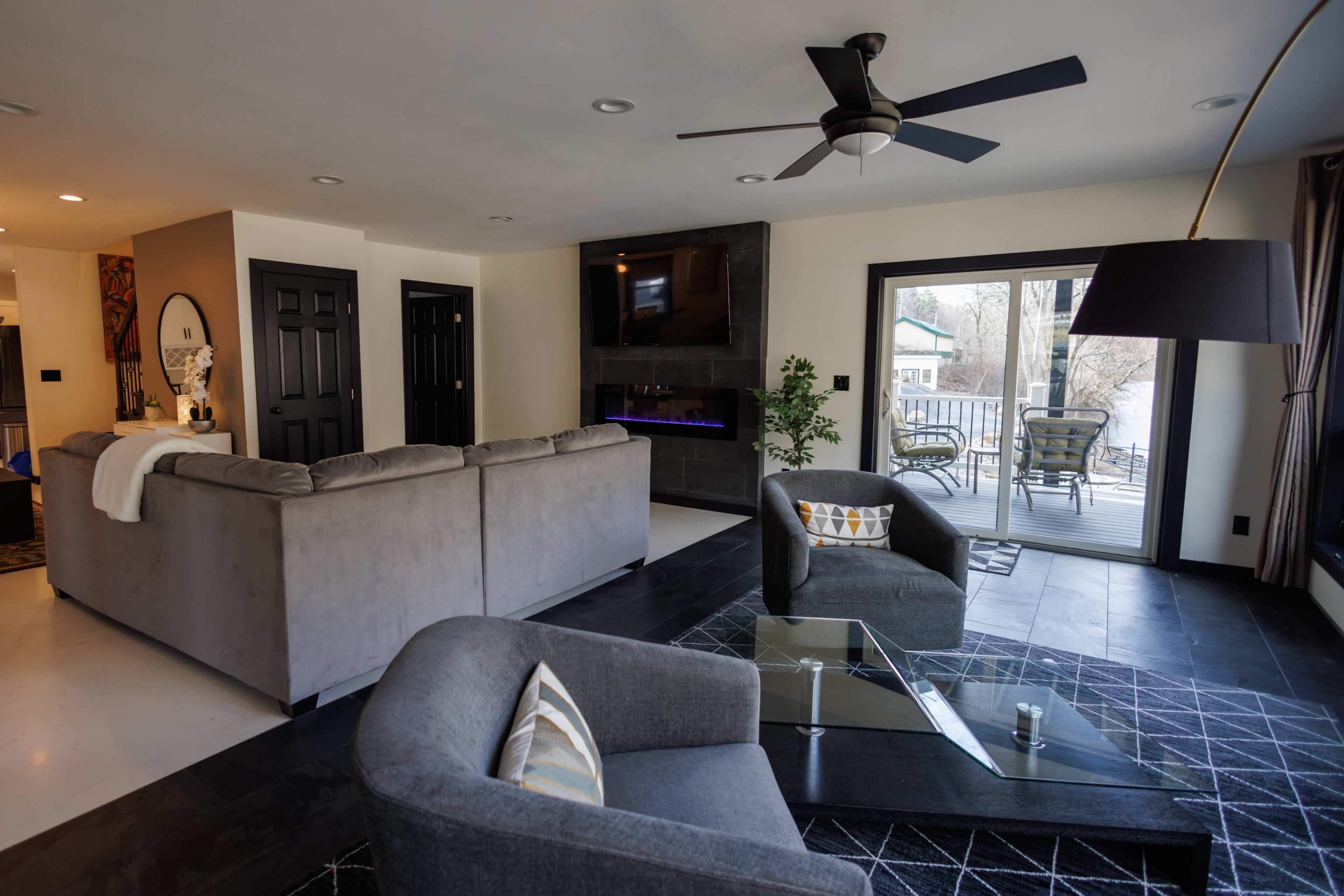 A modern living room featuring a gray sectional sofa, a glass coffee table, and a TV mounted above a fireplace, with large windows overlooking a deck.