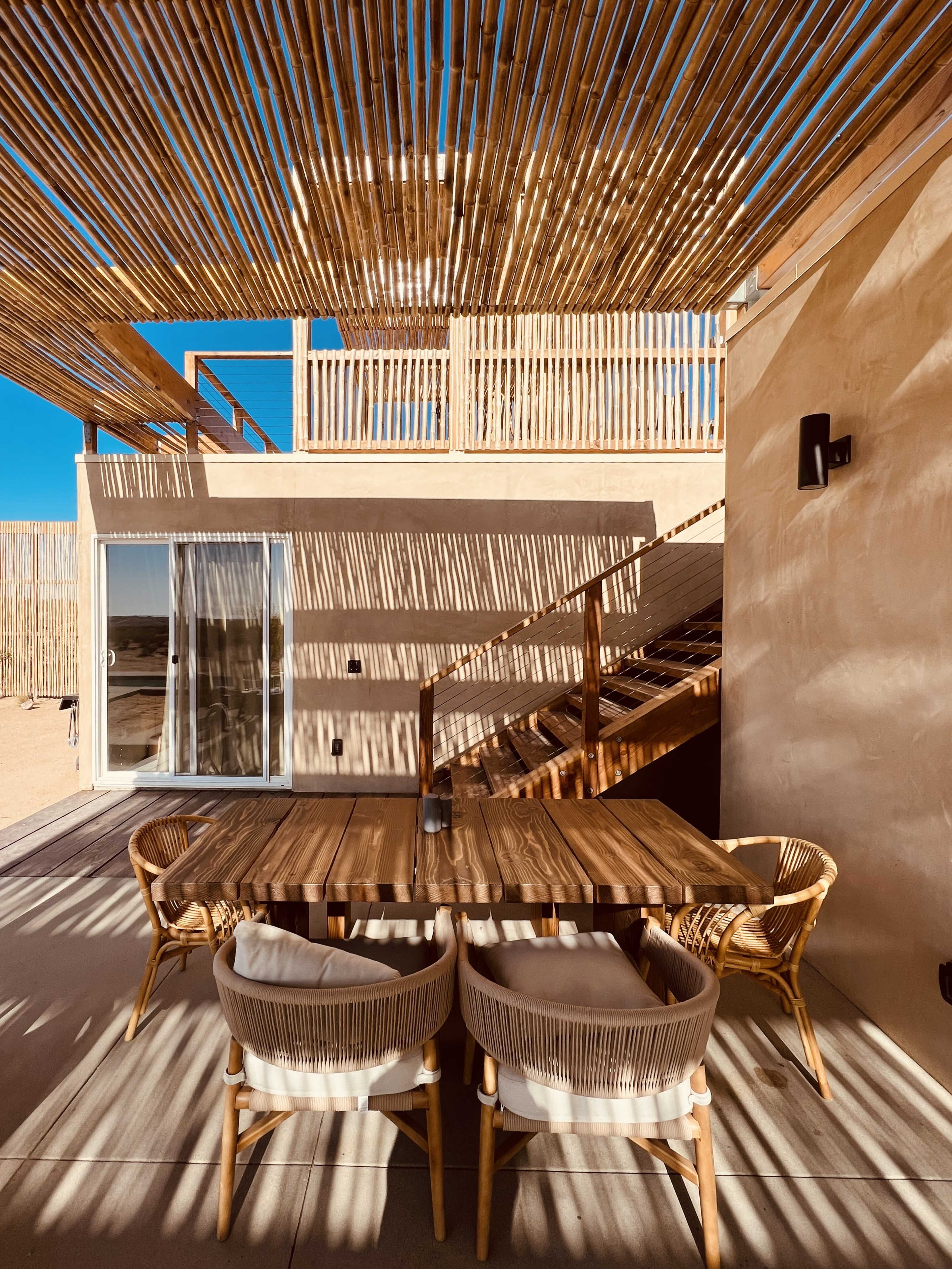 A modern outdoor dining area features a wooden table and chairs under a slatted overhead covering, with a staircase leading to a second level visible in the background.