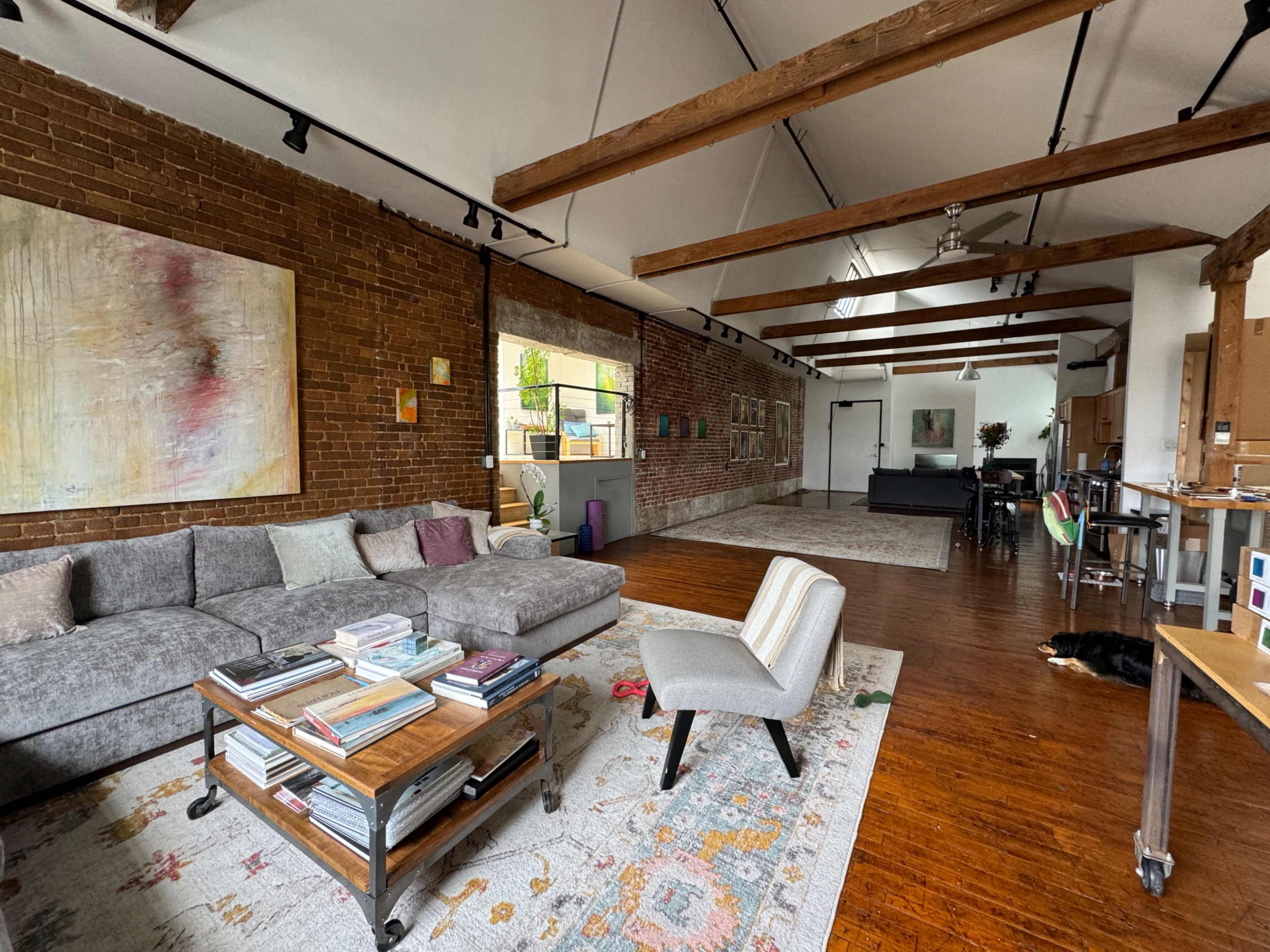 The image shows a spacious living area with brick walls, wooden beams, a gray couch, a coffee table filled with books, and a patterned rug, alongside a dining area and a dog lying on the floor.