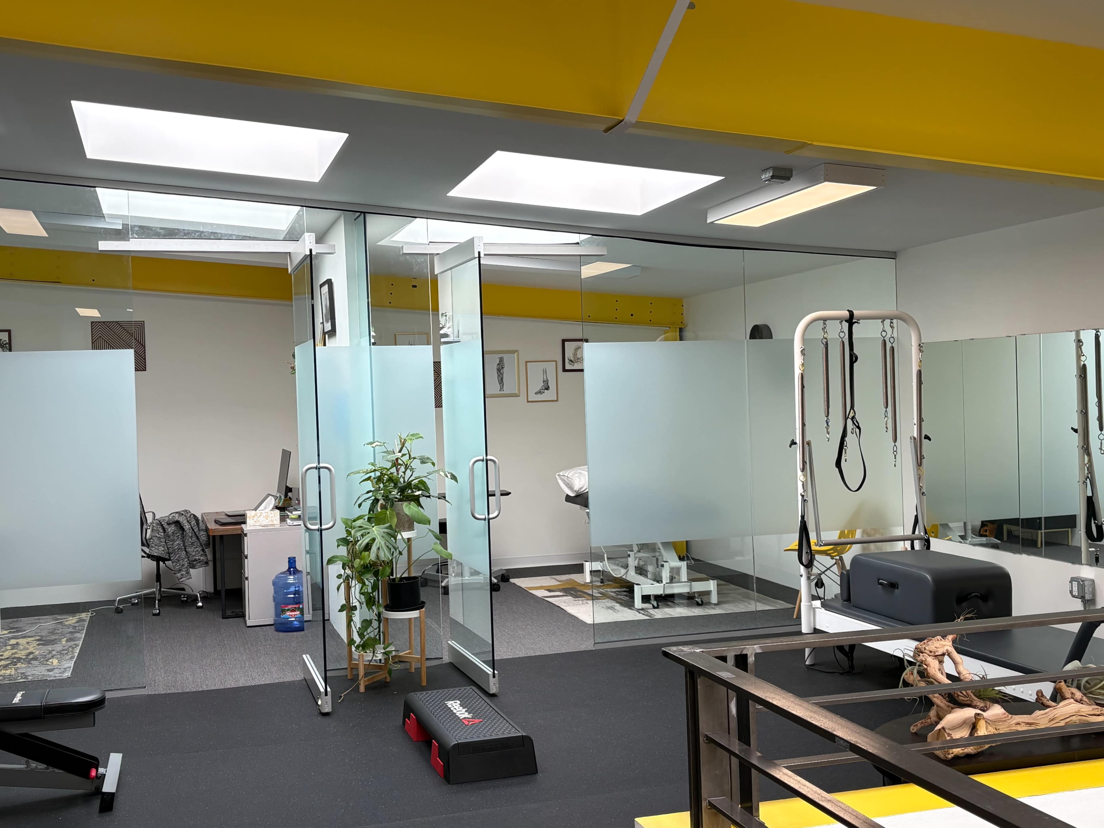 The image shows a modern gym or fitness studio with glass walls, fitness equipment, and an office area visible in the background.