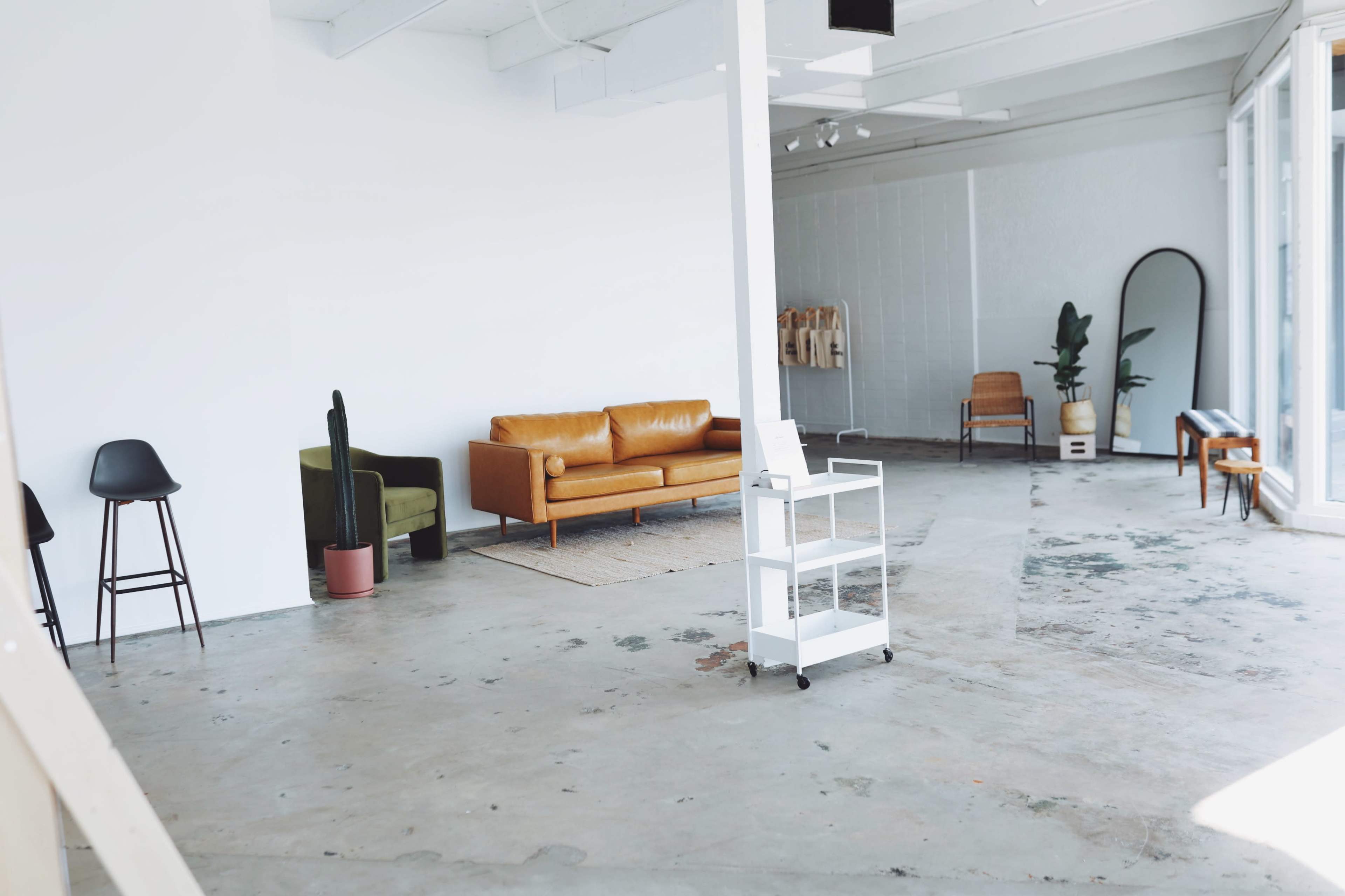 A spacious, minimalistic interior featuring a brown sofa, a green chair, a wooden chair, and a cart on wheels, with concrete flooring and white walls.