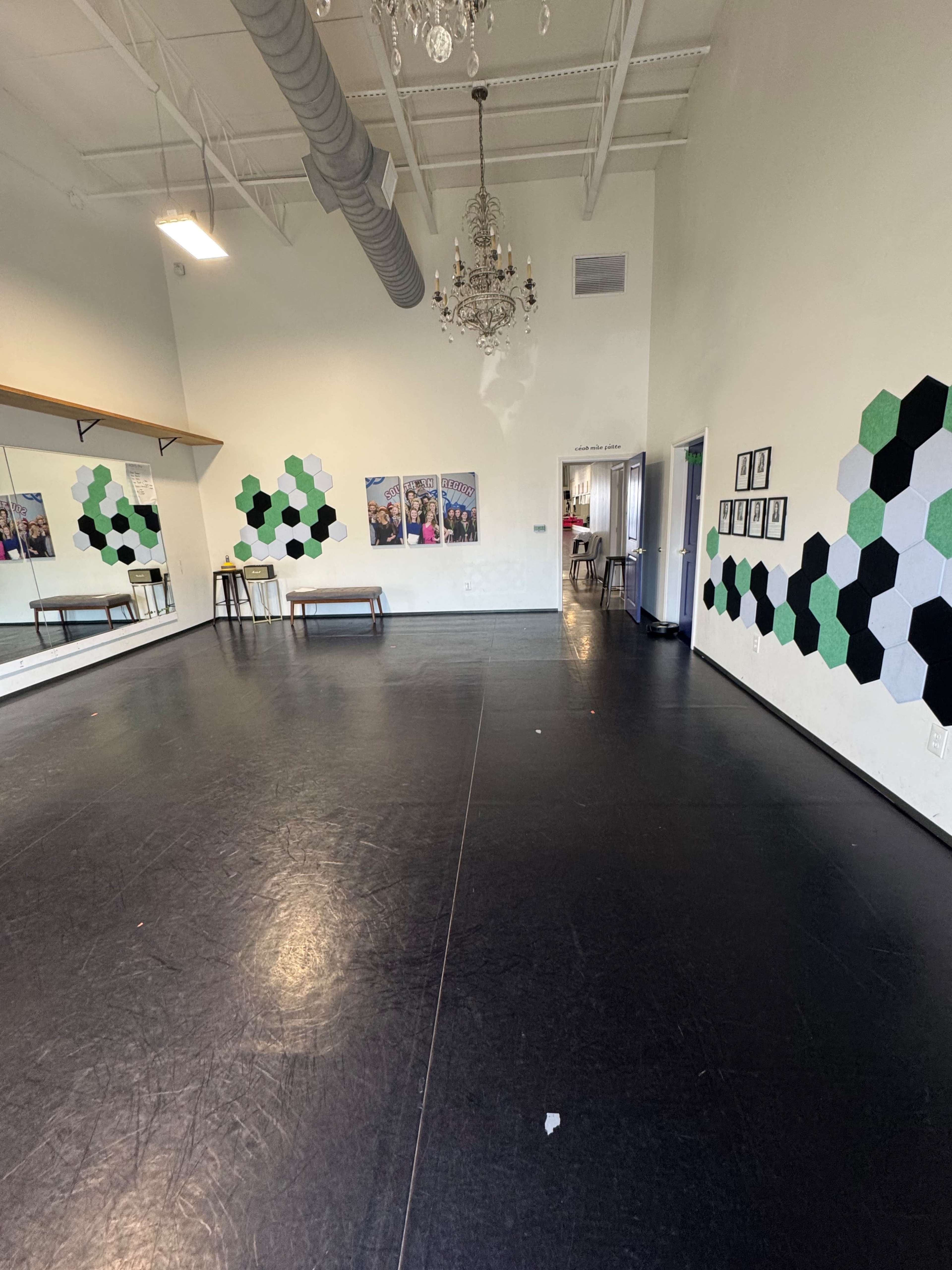 The image shows a spacious dance studio with a black floor, a mirrored wall, green and black hexagonal wall decorations, a chandelier, and several framed pictures on the walls.