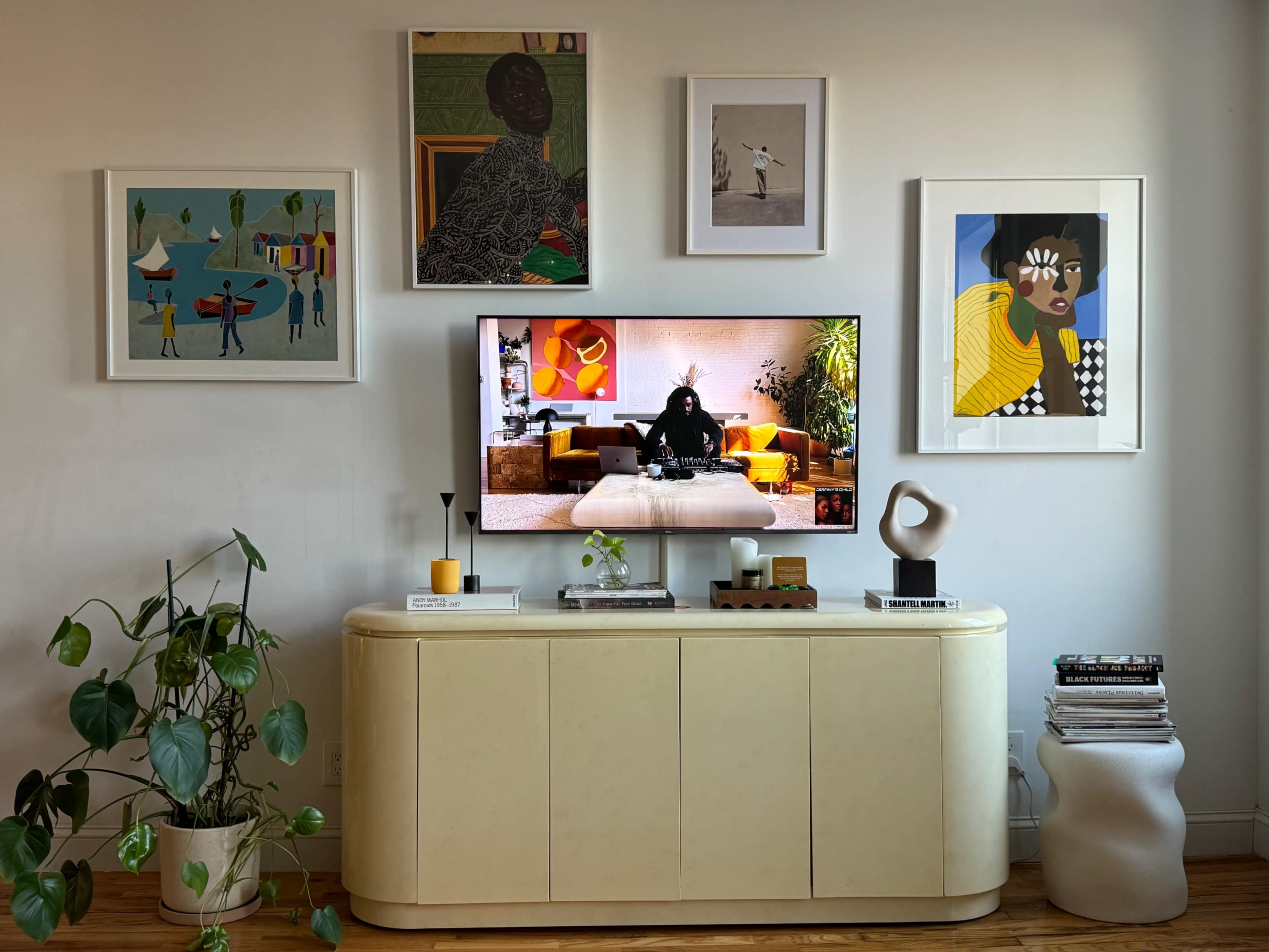 A modern living room features a wall adorned with framed artworks, a television displaying a scene on a light-colored media console, and a green plant in the foreground.