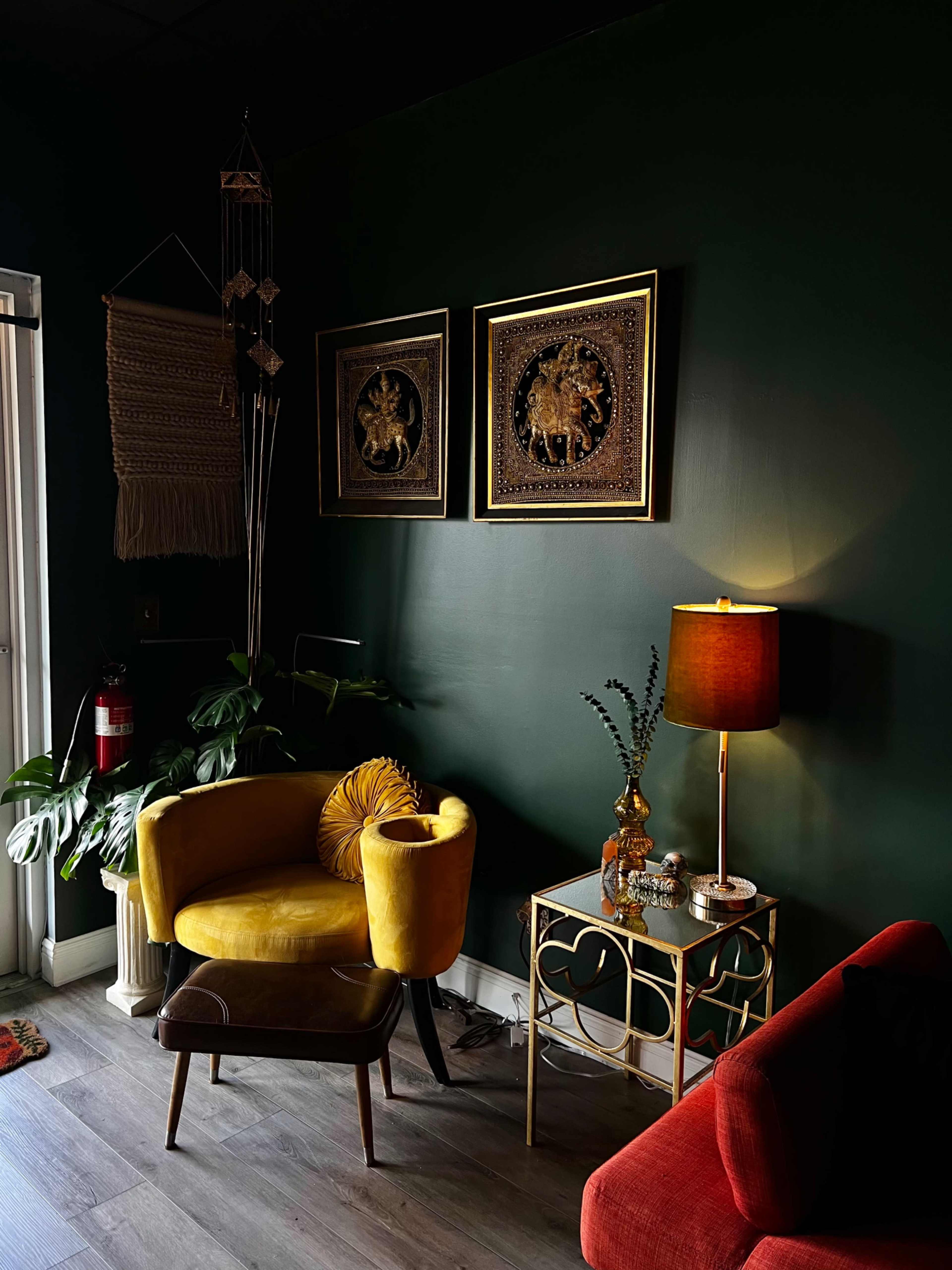 A yellow armchair and a red sofa sit in a dimly lit corner of a room with dark green walls, adorned with framed artwork and decorative plants.