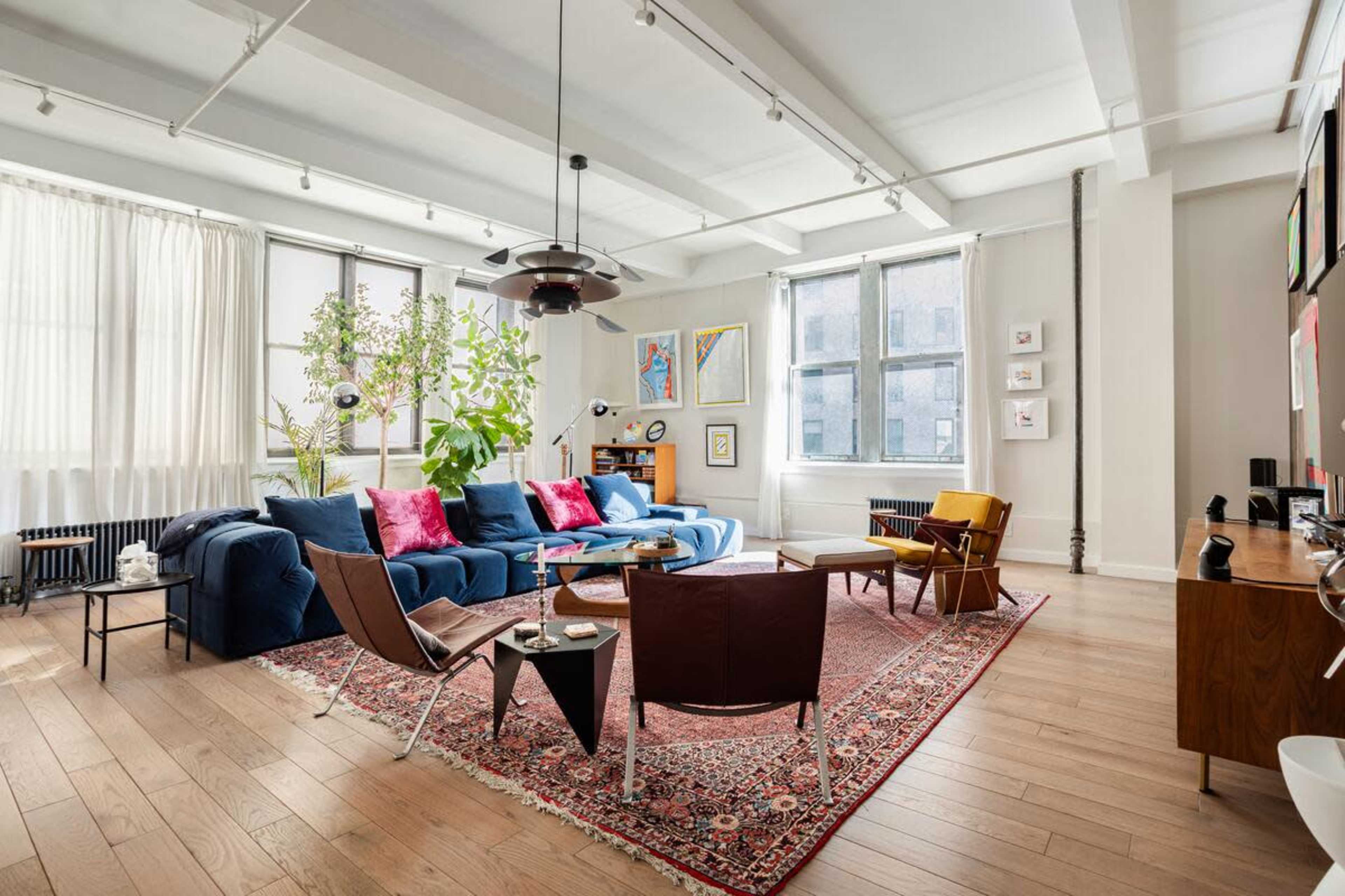 The image shows a spacious living room featuring a blue sofa with pink cushions, a large area rug, a wooden coffee table, and several plants near large windows.