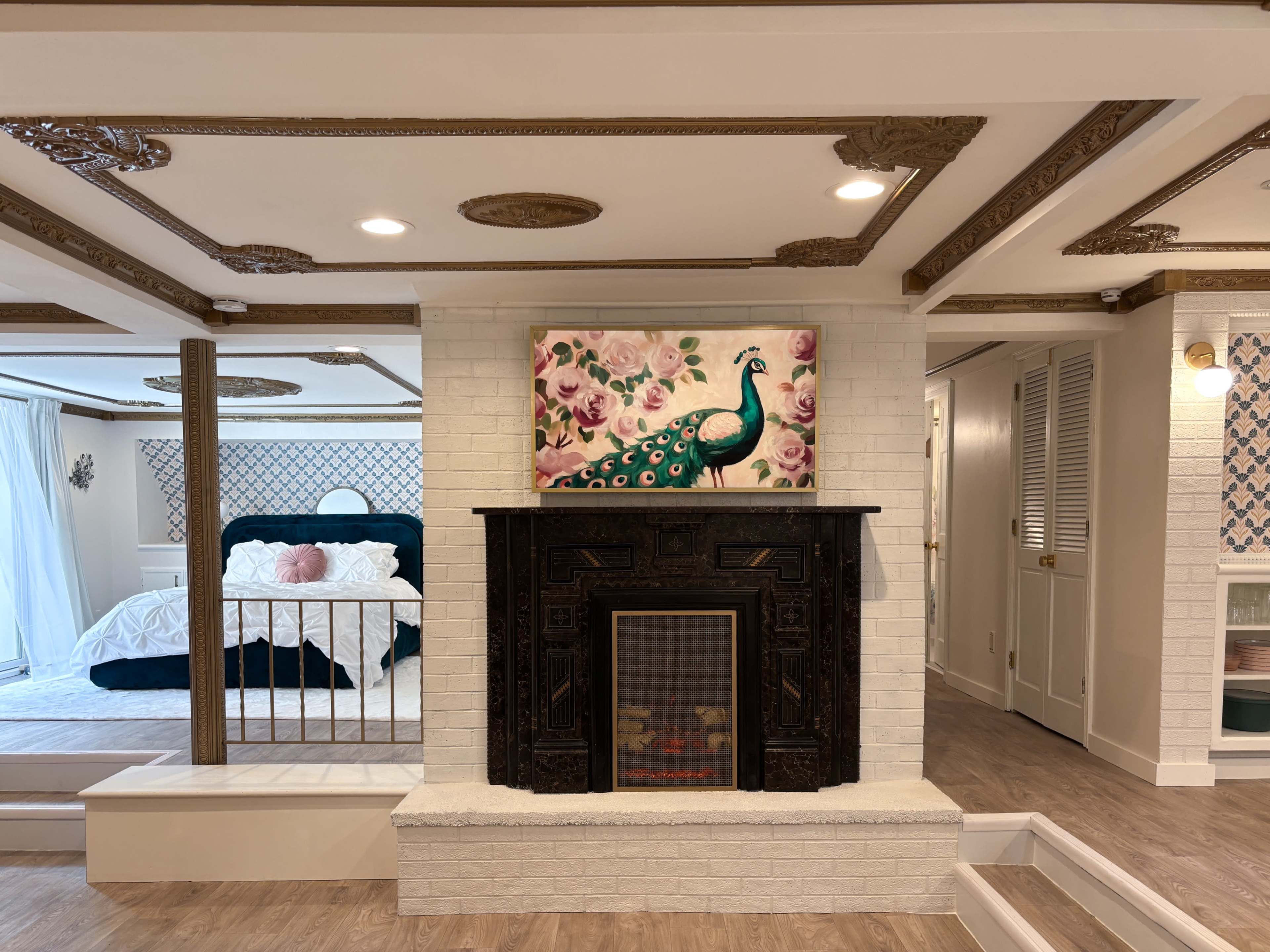 The image shows a cozy interior featuring a fireplace with a decorative mantel, a peacock painting above it, and a bedroom area with a large bed and light-colored furnishings in the background.