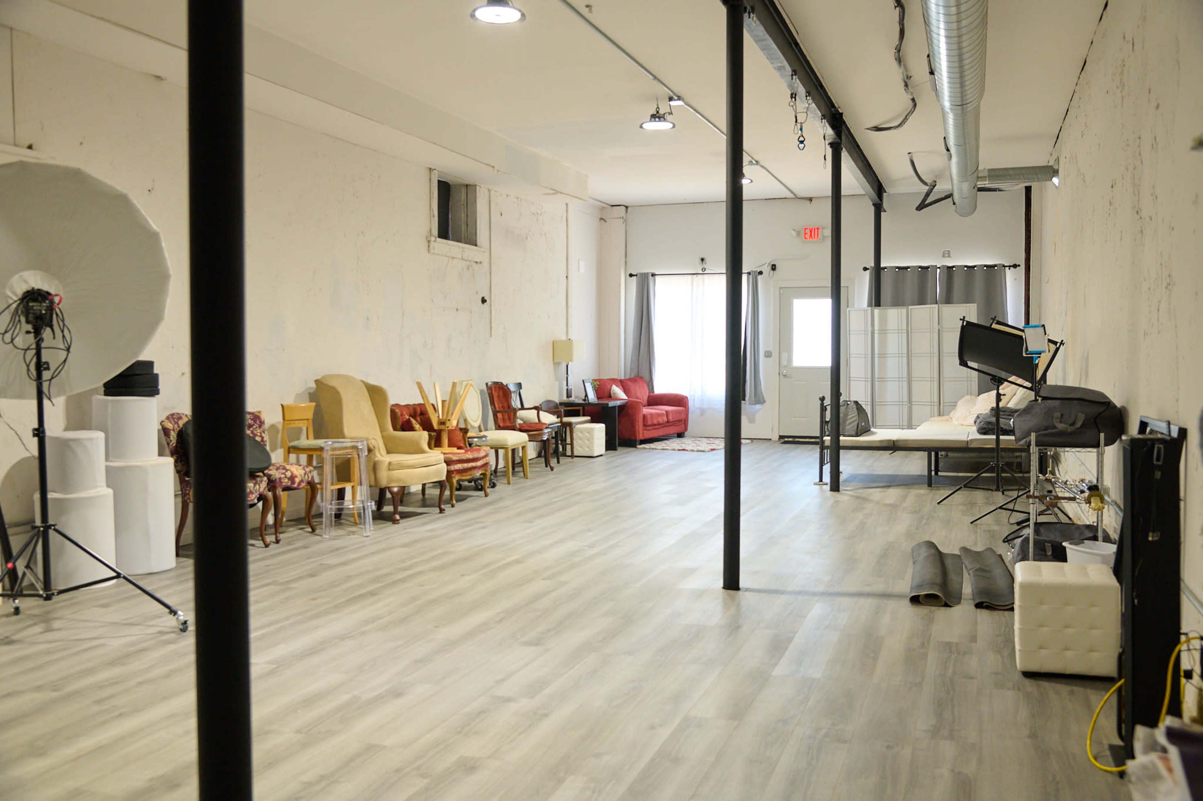 A spacious studio with various seating arrangements, including chairs and a sofa, along one side and a bed setup in the back corner.