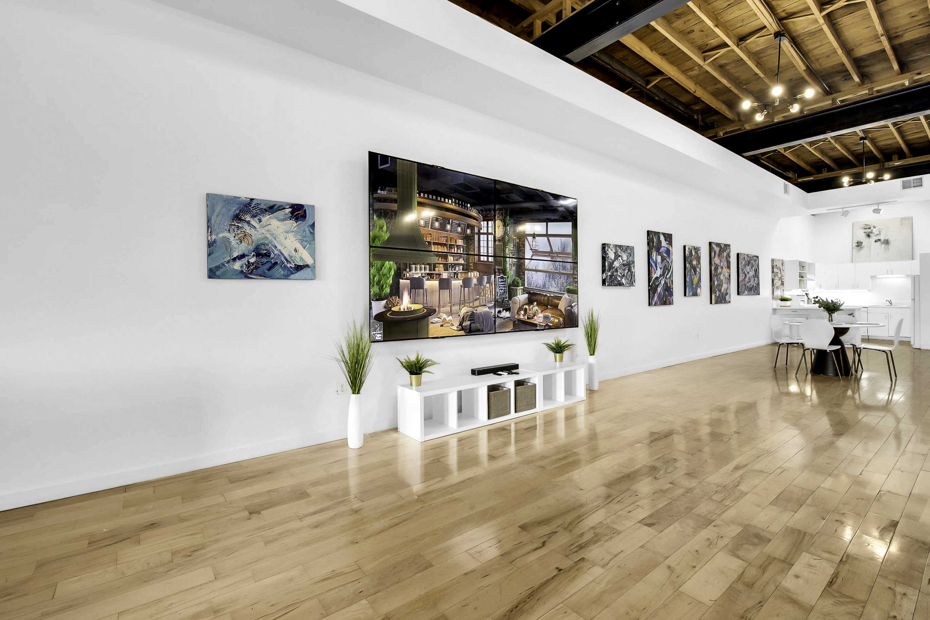 A modern gallery space featuring a large television display on a white wall, surrounded by various artworks and illuminated by overhead lighting.