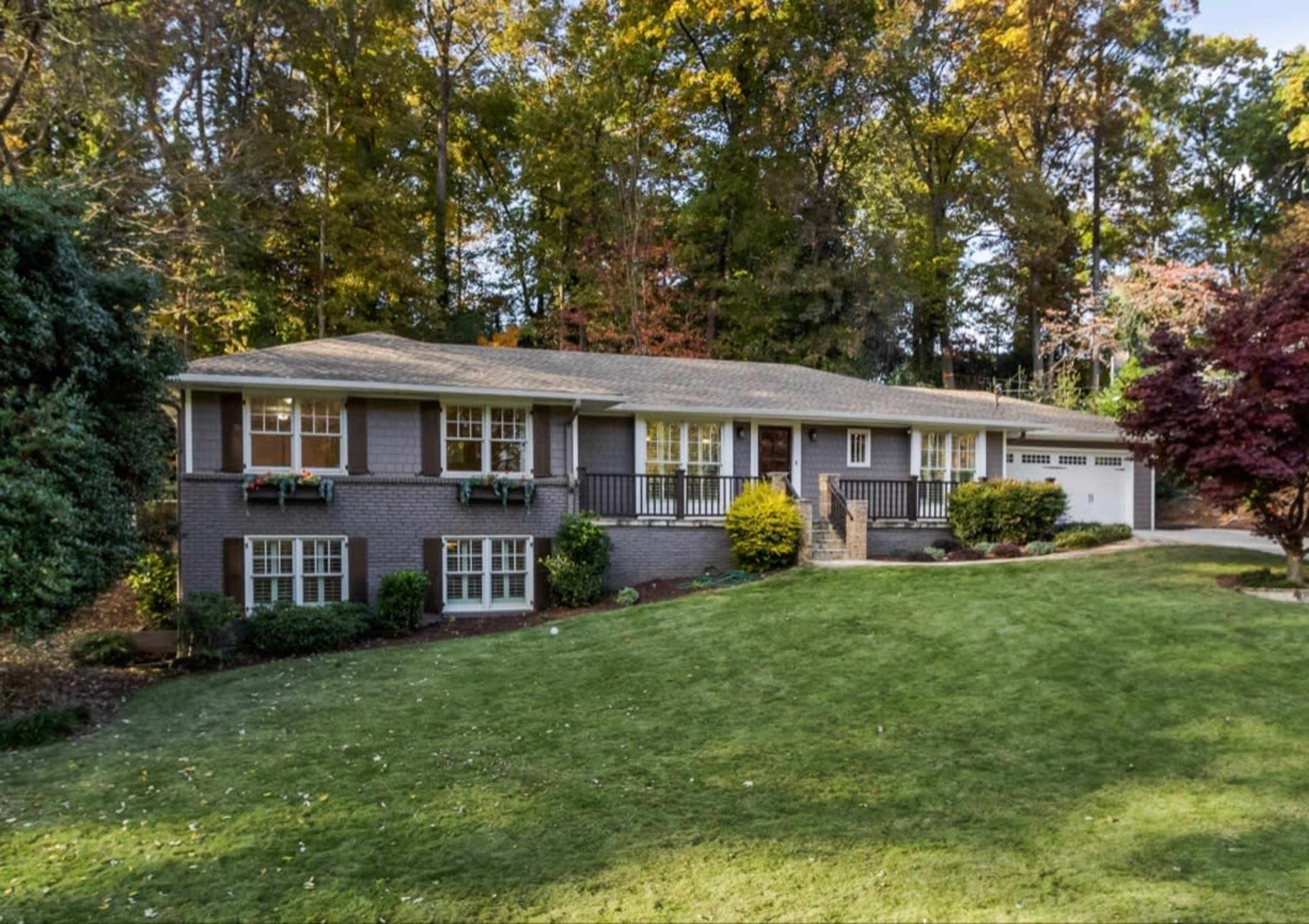 A split-level house with a gray brick facade and a landscaped yard is surrounded by trees with changing autumn foliage.