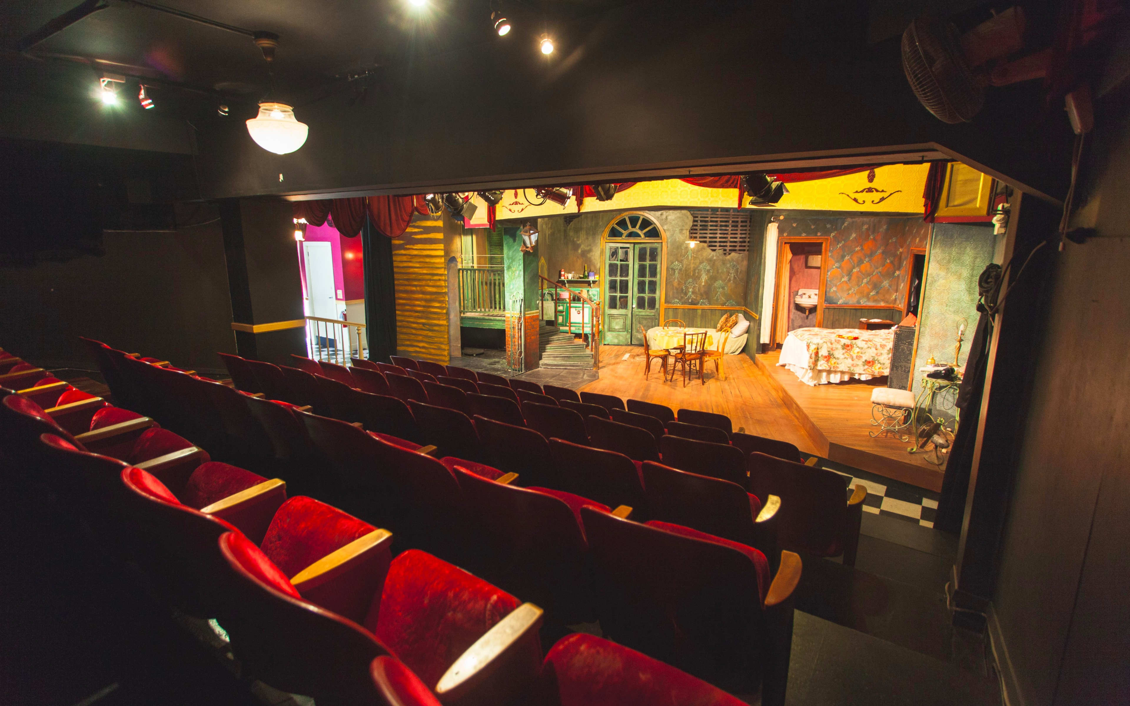 An empty theater with red seats facing a stage set with a wooden table, chairs, and a decorated backdrop.