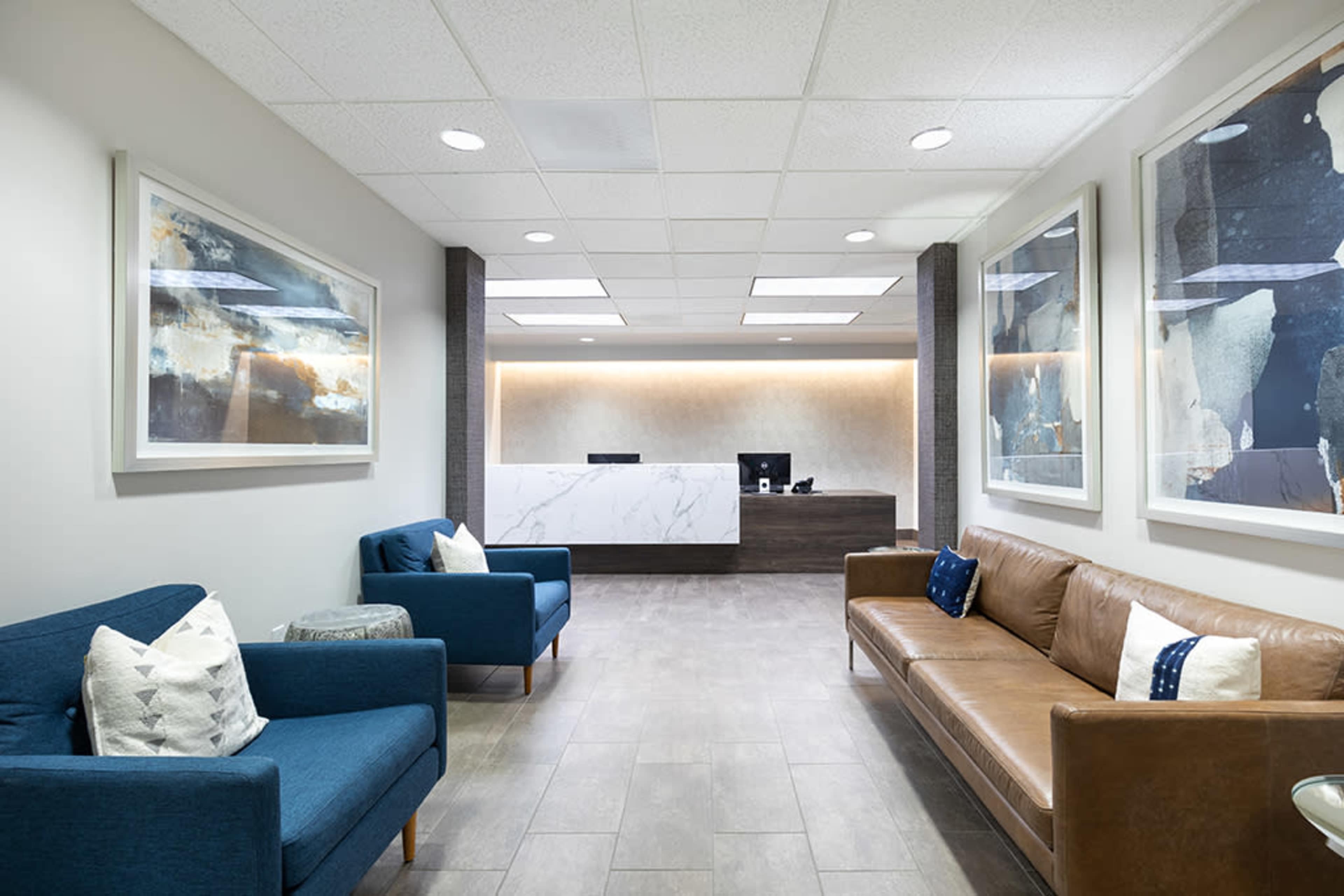 A modern reception area with a front desk, two blue couches, and a brown leather sofa, surrounded by large framed artworks and neutral-colored walls.