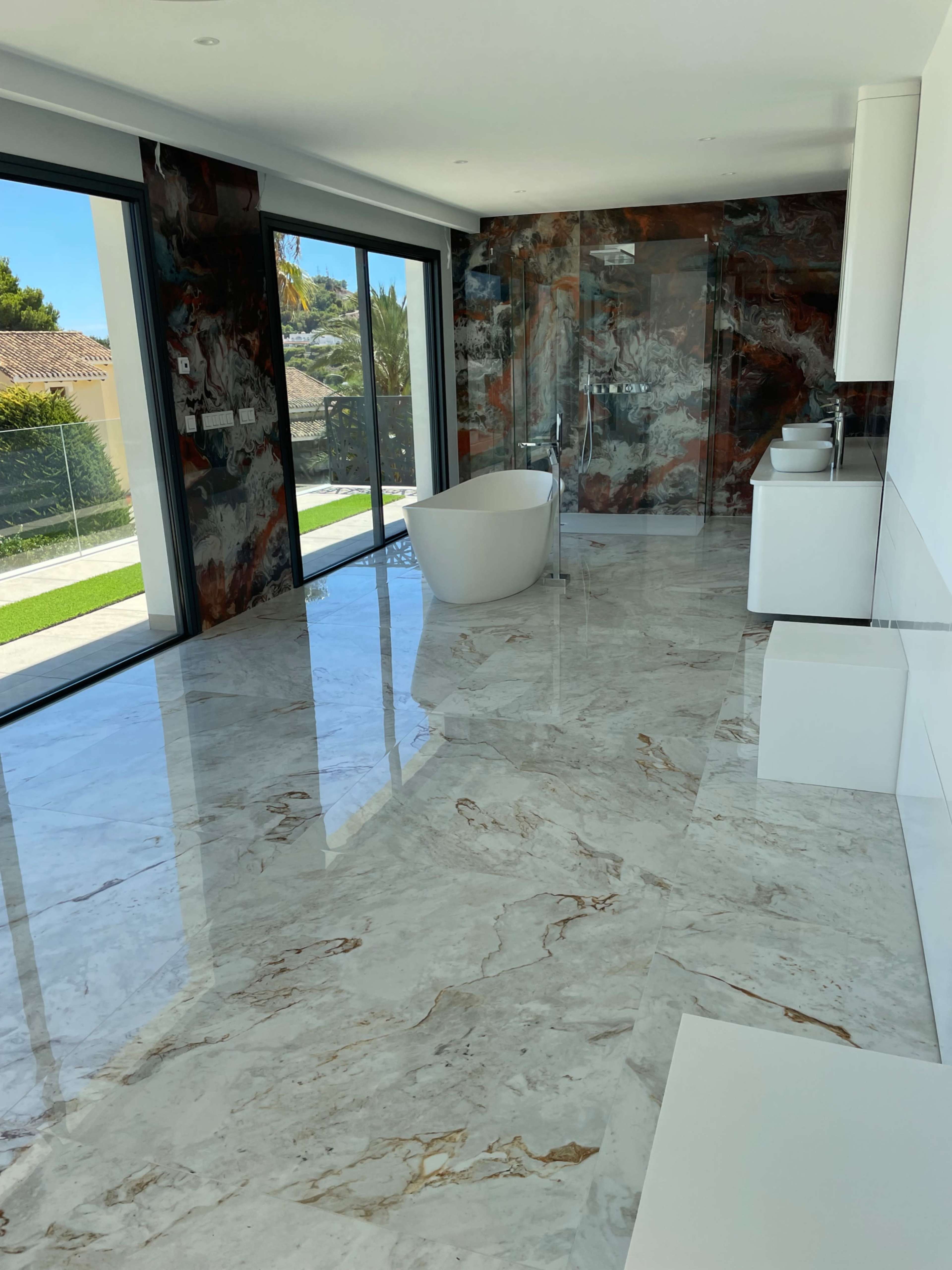 A spacious modern bathroom features a freestanding bathtub and large windows overlooking a landscaped exterior, with marble flooring and walls.