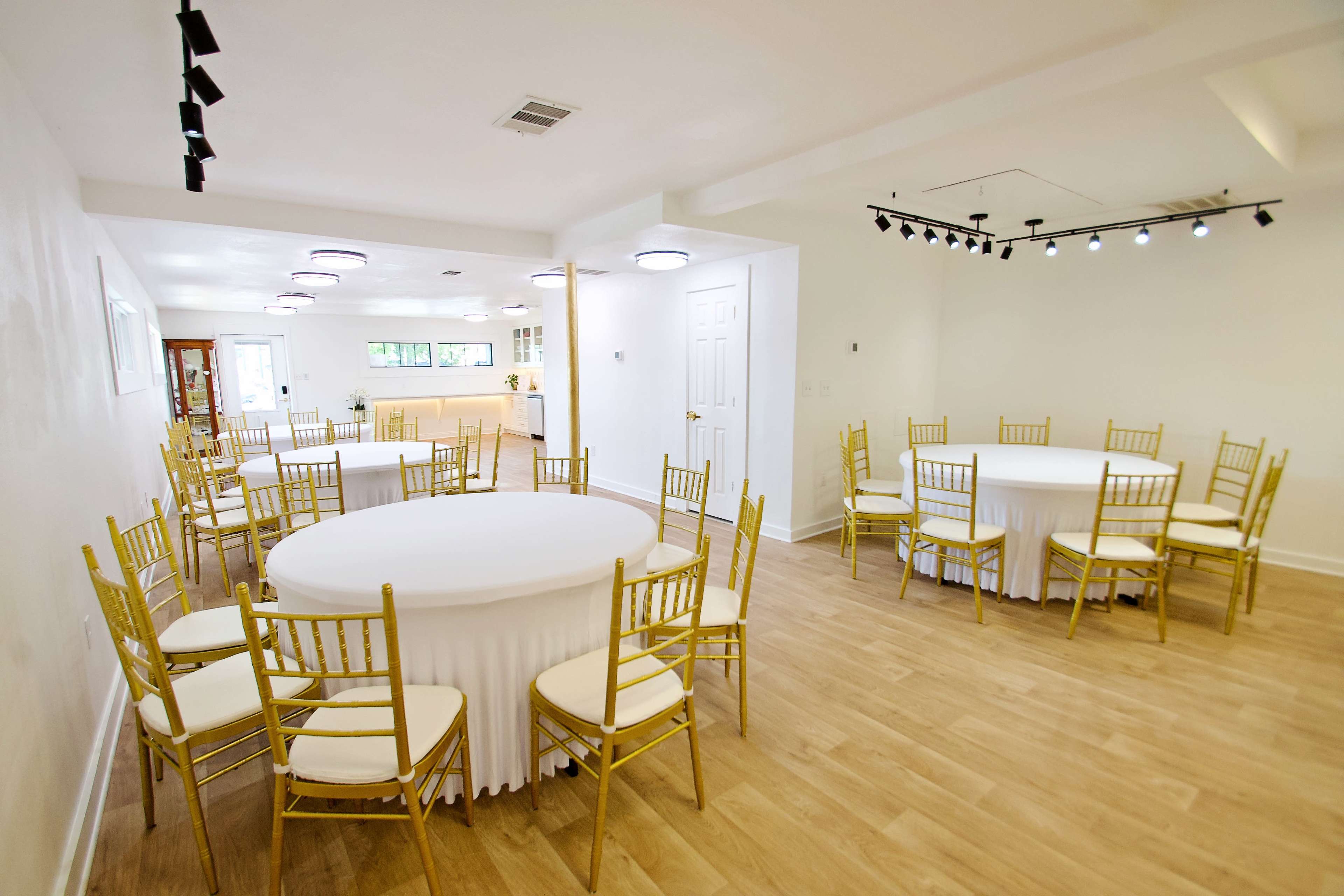 The image shows a spacious room arranged with several round tables covered in white tablecloths and surrounded by gold chairs.