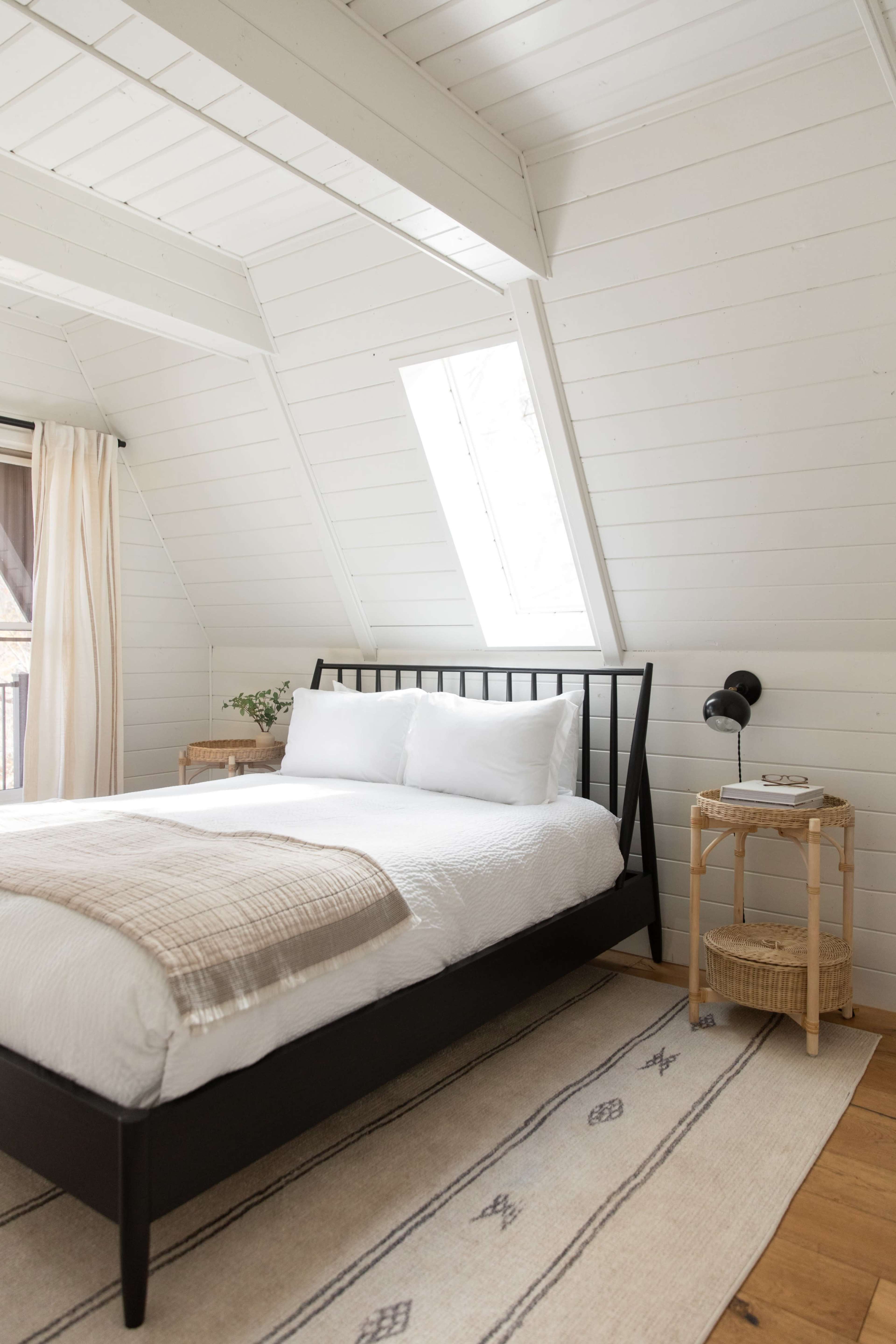 A minimalist bedroom features a black metal bed with white bedding, positioned under angled white wooden walls, accompanied by a nightstand and a woven rug.