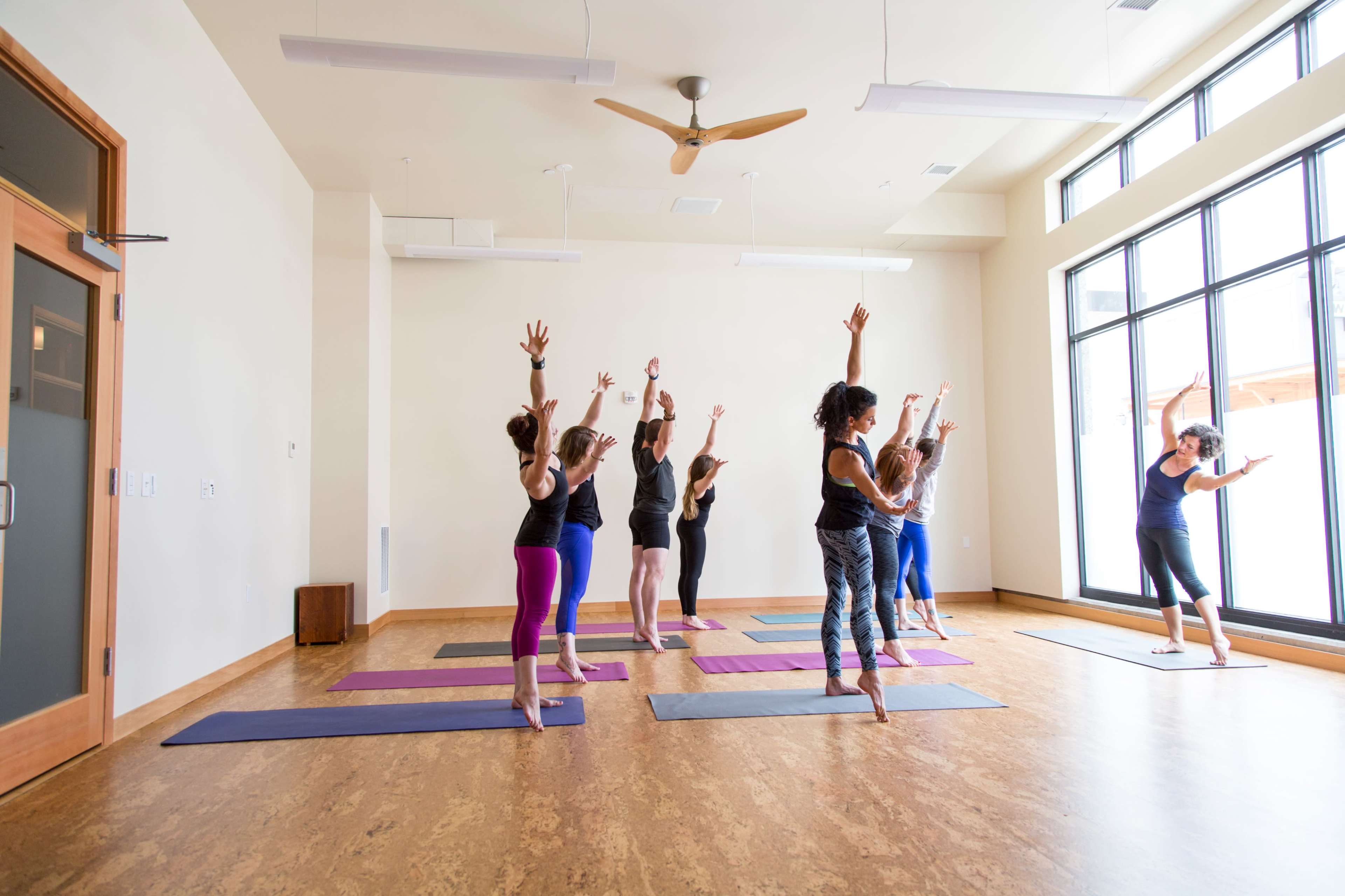 A group of people practices yoga in a spacious studio with large windows and wooden flooring.