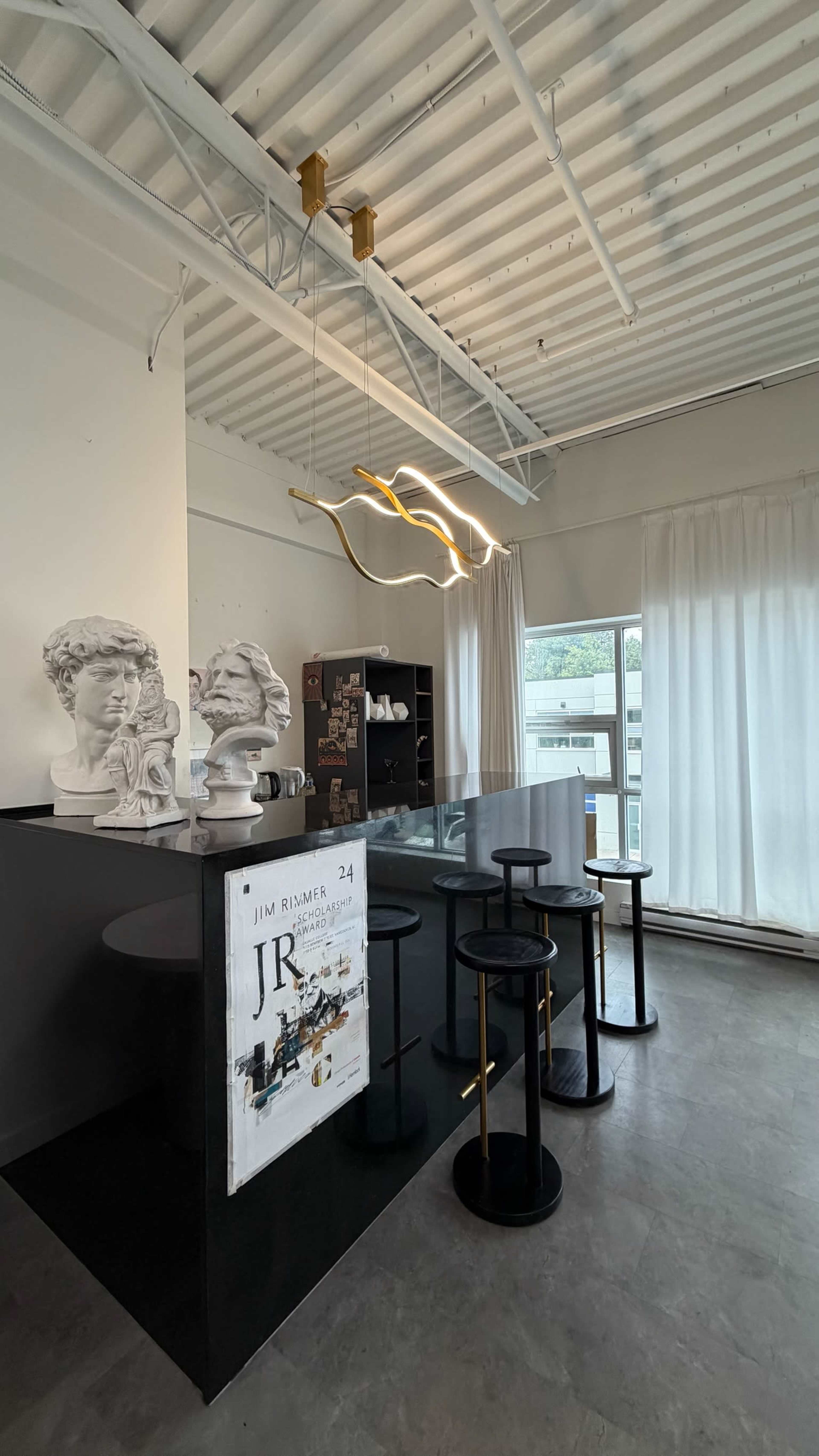 The image shows a modern coffee shop interior featuring a black counter, high stools, decorative busts on display, and large windows with white curtains.